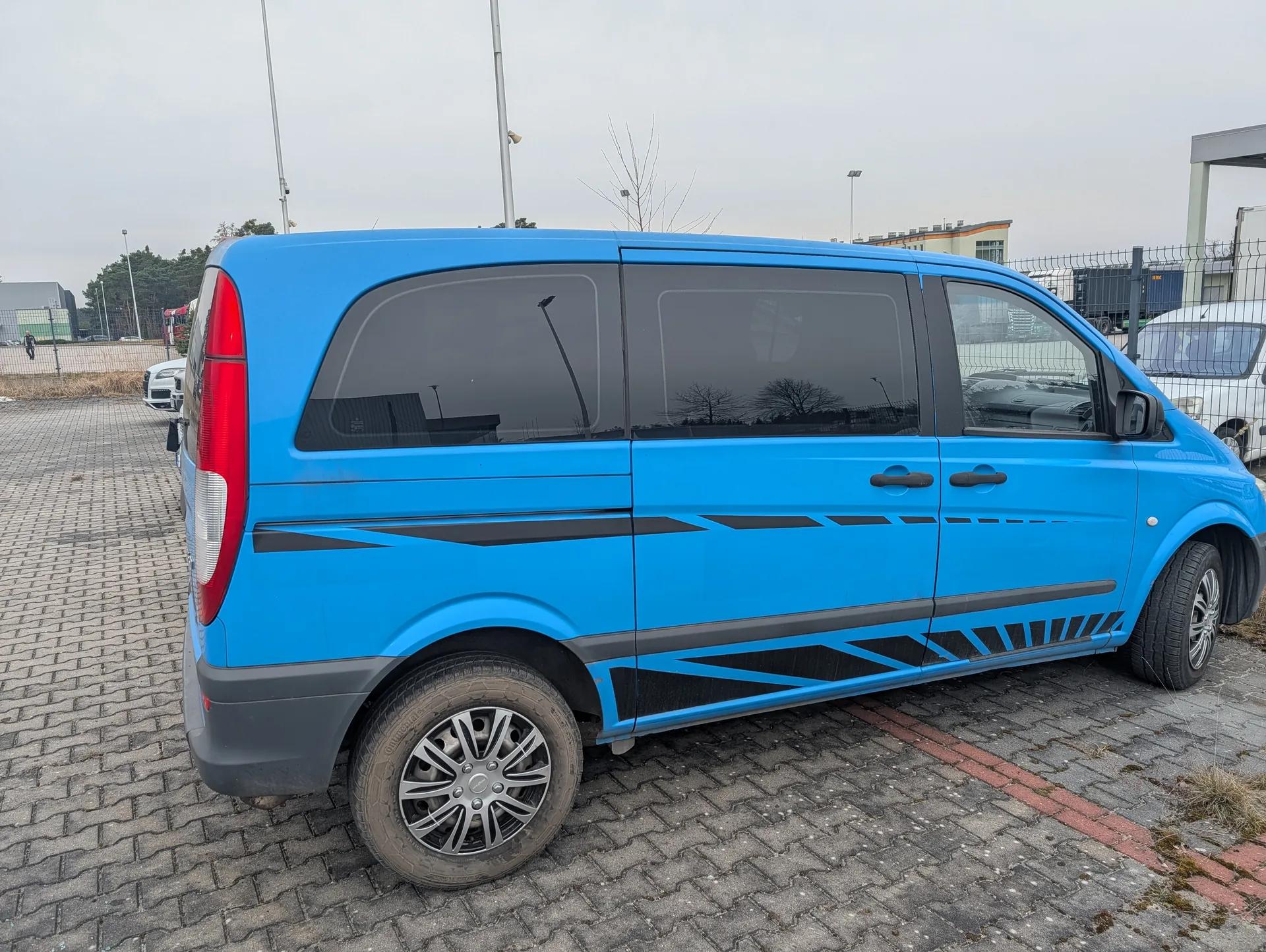 Niebieski furgon z czarnymi dekoracyjnymi pasami na bokach, zaparkowany na brukowanym placu. Furgon ma ciemne szyby, srebrne felgi i czerwone światło tylnego. W tle widoczne są ogrodzenie, inne pojazdy, budynki przemysłowe oraz drzewa bez liści. Niebo jest pochmurne.