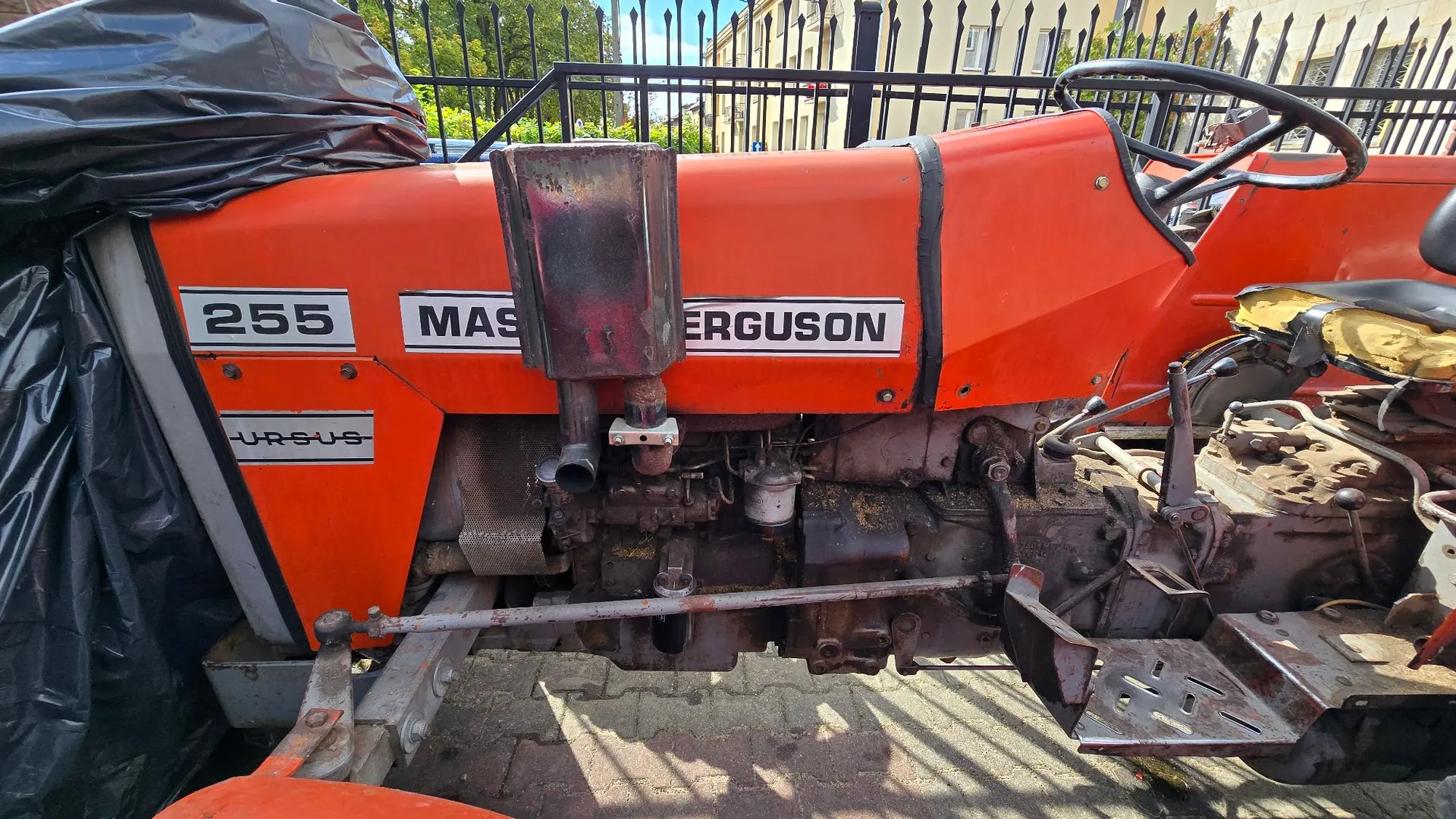 Zbliżenie na bok pomarańczowego traktora. Na masce widnieje biała tabliczka z napisem '255 MASSEY FERGUSON' oraz mniejszy napis 'URSUS' poniżej. Widoczny jest silnik, rura wydechowa oraz fragment kierownicy i siedzenia kierowcy. Przednia część traktora jest przykryta czarną folią.