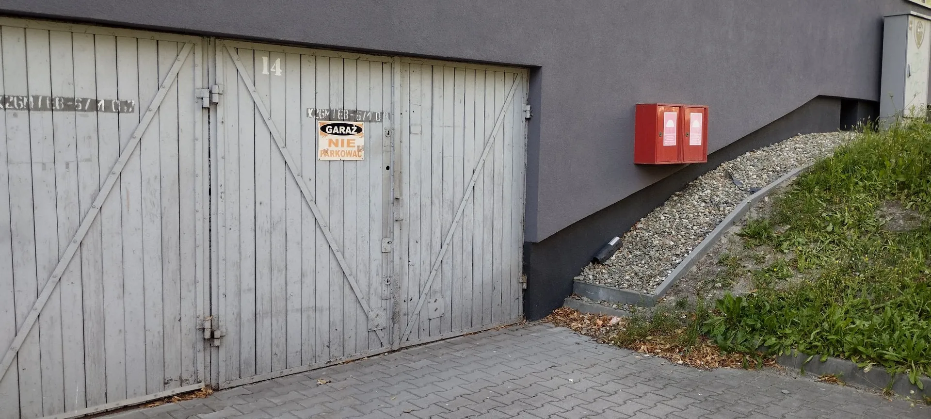Szare, drewniane drzwi garażowe z numerem 14 nad wejściem. Na drzwiach znajduje się biała tabliczka z napisem „GARAŻ NIE PARKOWAĆ”. Obok garażu znajduje się szara ściana budynku, na której zamontowano czerwoną skrzynkę przeciwpożarową. Po prawej stronie widoczny jest nasyp z kamieni i zielona roślinność.