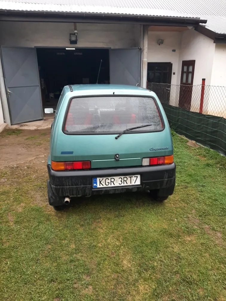 Z tyłu widoczny zielony samochód osobowy z tablicą rejestracyjną KGR 3RT7 (PL) na czarnym zderzaku. Samochód stoi na trawie przed budynkiem z otwartymi szarymi bramami garażowymi. W tle biały budynek z metalowym dachem, ogrodzenie siatkowe z zielonym tkaninowym osłonem oraz drzwi i okno z brązowymi ramami. Na tylnej szybie samochodu widoczny wycieraczka, a na bagażniku niewyraźny napis. Podwozie i zderzak pokryte są błotem.