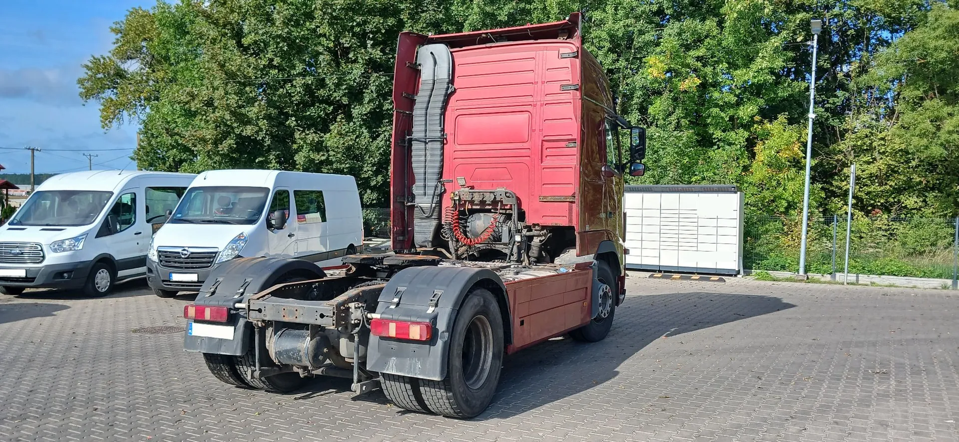 Czerwona ciągnik siodłowy bez naczepy, zaparkowany na kostce brukowej. Pojazd jest widoczny z tyłu i z boku, ukazując ramę i układ napędowy. W tle znajdują się dwa białe samochody dostawcze, biały kontener oraz gęsta ściana zielonych drzew pod niebieskim niebem.