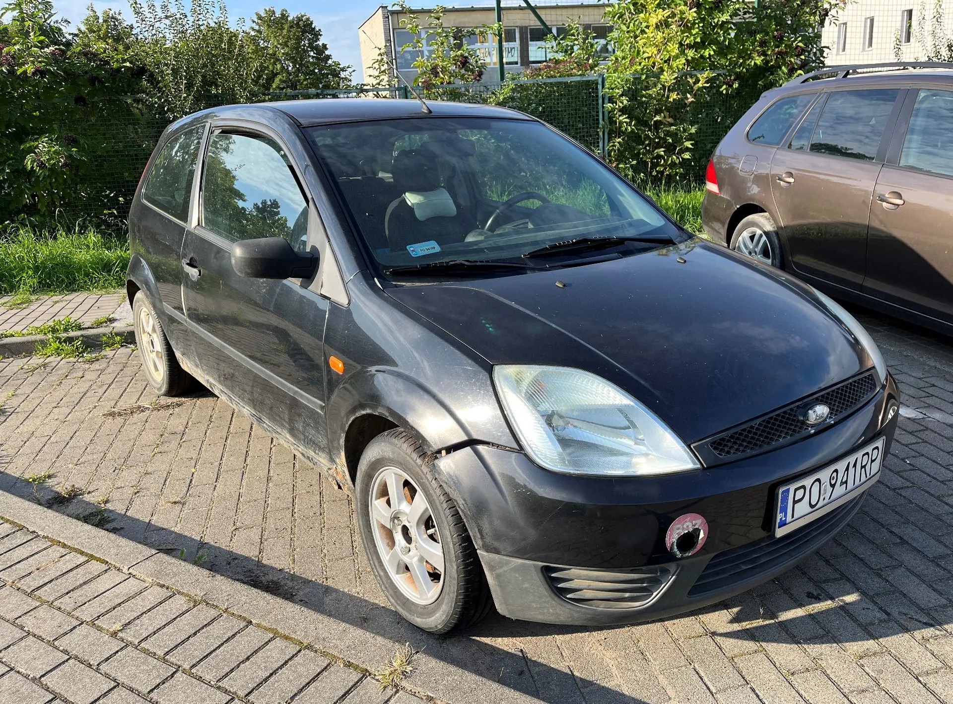 Czarny, trzy drzwiowy samochód osobowy marki Ford Fiesta zaparkowany na kostce brukowej. Pojazd ma polską tablicę rejestracyjną o numerze PO 941RP. W tle widoczna jest zieleń, krzewy oraz fragment jasnego budynku.