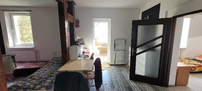 Wnętrze mieszkania z widocznym pokojem. Po lewej stronie okno z widokiem na drzewa, pod nim grzejnik i łóżko z kolorową narzutą. W centrum stół z krzesłem, na którym leży ciemna kurtka. Przy stole stoi osoba w ciemnej sukience, odwrócona tyłem. W tle drzwi prowadzące do innego pomieszczenia z widocznym krzesłem. Po prawej stronie metalowy stojak oraz szklane drzwi z ciemną ramą prowadzące do pokoju z łóżkiem o kolorowej pościeli. Podłoga wyłożona dywanem w geometryczny wzór, ściany jasne. Brak czytelnego tekstu.