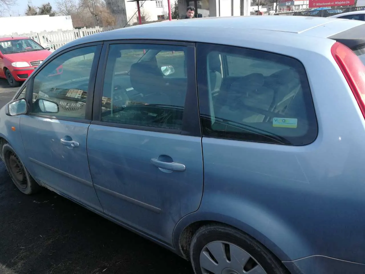 Niebieski samochód osobowy typu minivan zaparkowany na asfaltowej powierzchni. Widoczne są boczne drzwi, przednie i tylne koła, lusterka oraz szyby. Na tylnej szybie naklejka z napisem "V O". W tle czerwony samochód, biały płot, budynki i osoba stojąca za pojazdem. Tło obejmuje również drzewa i jasne niebo.