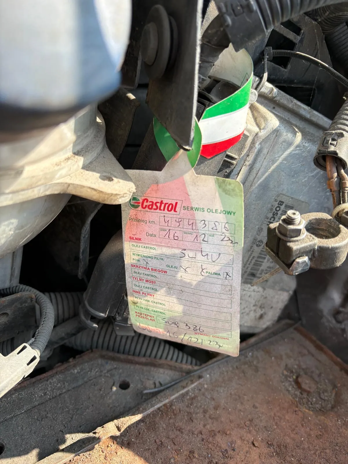 Zbliżenie na naklejkę Castrol SERWIS OLEJOWY przyklejoną do silnika samochodu. Naklejka zawiera informacje: Przebieg km: 494386, Data: 16 12 23, oraz sekcje SILNIK, OLEJ CASTROL, WYMIENIONO FILTR, SKRZYNIA BIEGÓW, TYLNY MOST, INNE PŁYNY. Widoczne są ręcznie wpisane dane, w tym 3u4v oraz zaznaczenia X. Obok naklejki znajduje się wstążka w kolorach zielonym, białym i czerwonym. W tle widoczne są elementy silnika, w tym czarna rura, metalowe części i etykieta z napisem BOSCH. Na dole obrazu widoczna jest rdzewna powierzchnia.