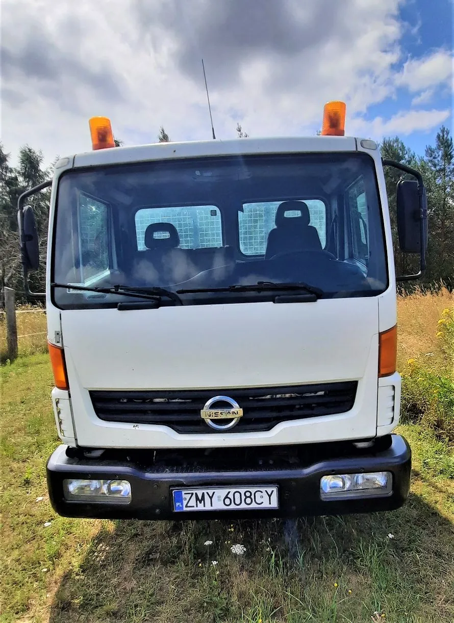 Widok z przodu białego samochodu ciężarowego marki Nissan. Pojazd posiada dwa pomarańczowe koguty na dachu oraz czarny zderzak z wbudowanymi reflektorami. Tablica rejestracyjna samochodu to ZMY 608CY. Auto stoi na trawiastym terenie na tle drzew i zachmurzonego nieba.