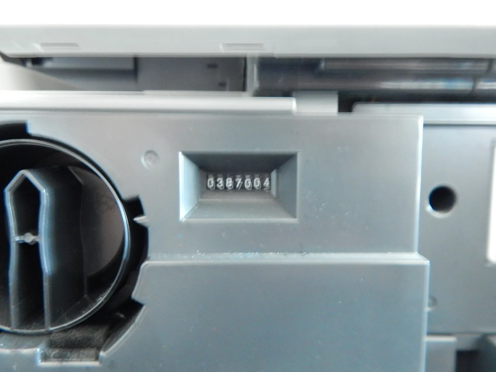 Zbliżenie na szary, plastikowy element urządzenia technicznego. W centralnej części znajduje się wnęka z mechanicznym licznikiem wyświetlającym numer 0387004. Po lewej stronie widoczny jest fragment czarnego, okrągłego elementu z plastikowymi żłobieniami.