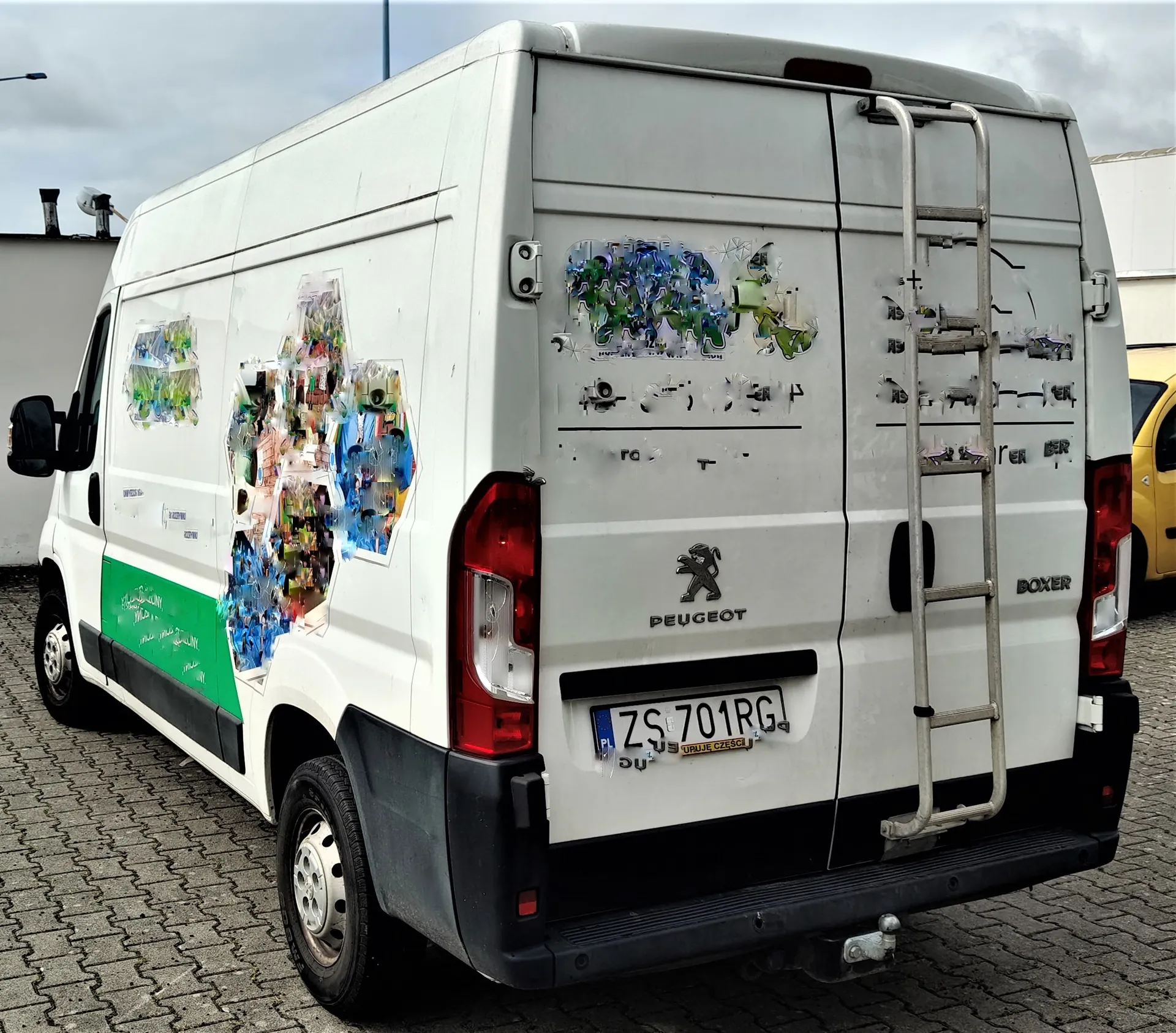 Biały furgon marki Peugeot Boxer zaparkowany na kostce brukowej. Pojazd ma zielony pas na dolnej części boku. Na tylnych drzwiach oraz boku samochodu widoczne są pozostałości po zdrapanych naklejkach i grafikach. Na tylnych drzwiach znajduje się logo Peugeot, napis BOXER oraz tablica rejestracyjna o numerze ZS 701RG. Z tyłu pojazdu zamontowana jest drabinka.