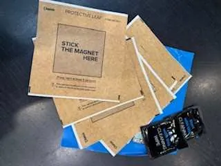 Kilka brązowych kartonowych kopert lub opakowań ułożonych na niebieskiej powierzchni. Na opakowaniach widoczny tekst: 'STICK THE MAGNET HERE' oraz 'PROTECTIVE FILM'. W lewym górnym rogu znajduje się czarna paczka. Tło stanowi ciemna powierzchnia.