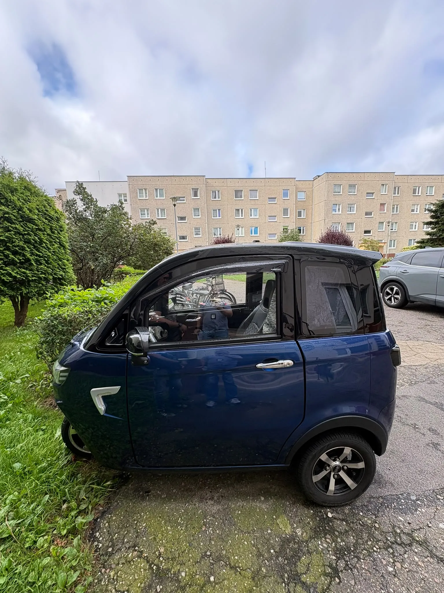 Niebieski, kompaktowy samochód elektryczny zaparkowany na poboczu obok trawnika. W tle widoczny jest wielopiętrowy blok mieszkalny z jasnej cegły oraz fragment innego, srebrnego samochodu. Niebo jest zachmurzone.