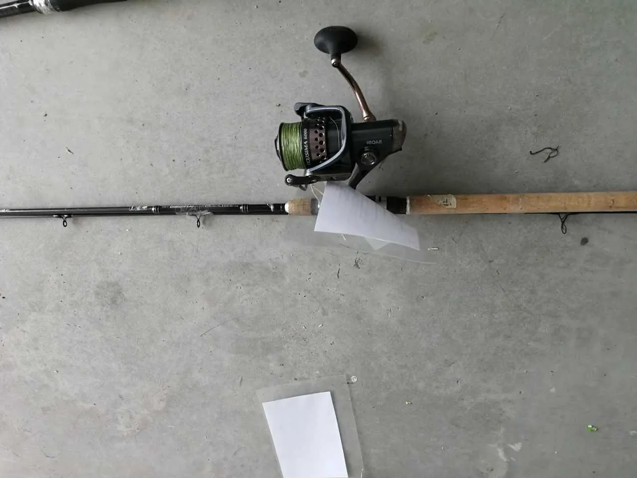 Horyzontalnie ułożony wędkarski kij z przewodnicą. Na przewodnicy widoczny zielony sznurek, a na niej napisy 'COSMOS 6000' oraz '100X'. Pod przewodnicą znajduje się biały arkusz papieru, a poniżej kija kolejny biały arkusz w przezroczystej folii. Tło stanowi szara powierzchnia betonowa z drobnymi zabrudzeniami.