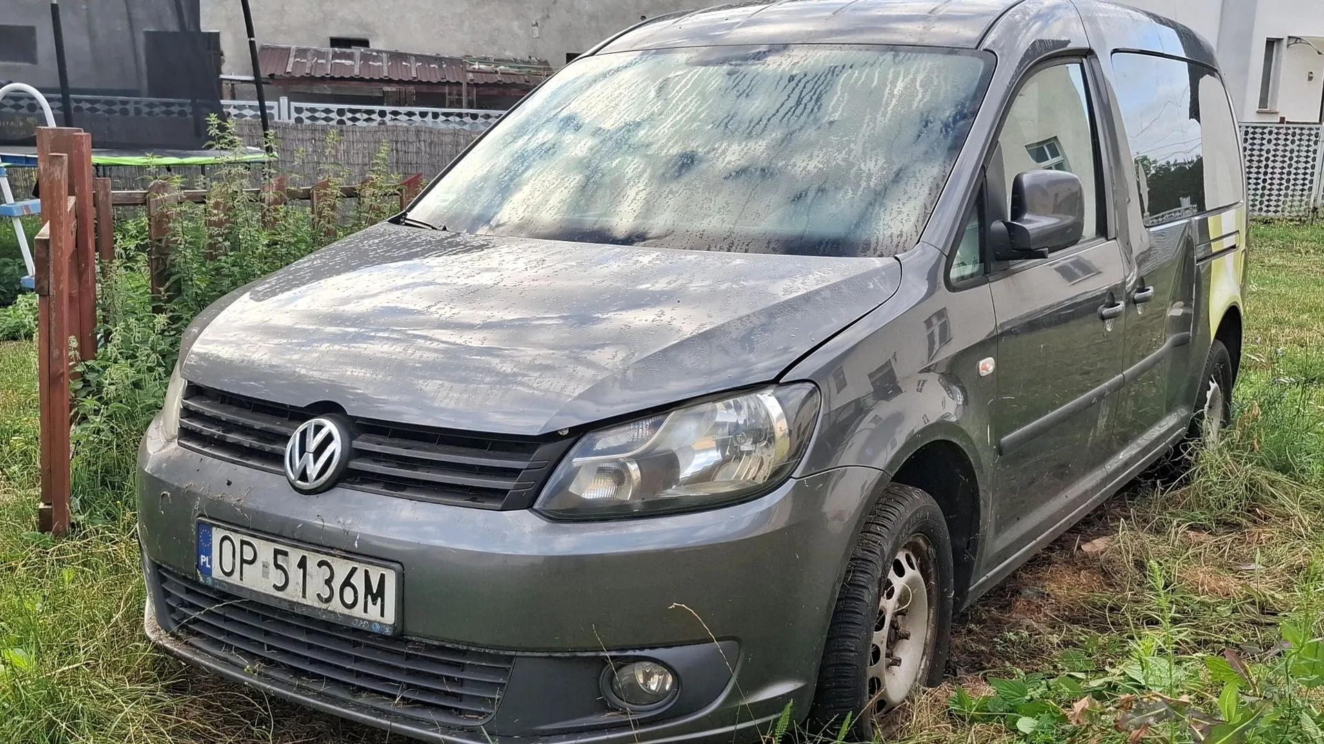 Szary samochód dostawczy marki Volkswagen zaparkowany na trawiastym terenie. Pojazd jest brudny, z widocznymi kroplami wody na przedniej szybie i masce. Na przodzie znajduje się tablica rejestracyjna o numerze OP 5136M. W tle widoczne są niskie budynki, ogrodzenie z drewnianych palików oraz roślinność.
