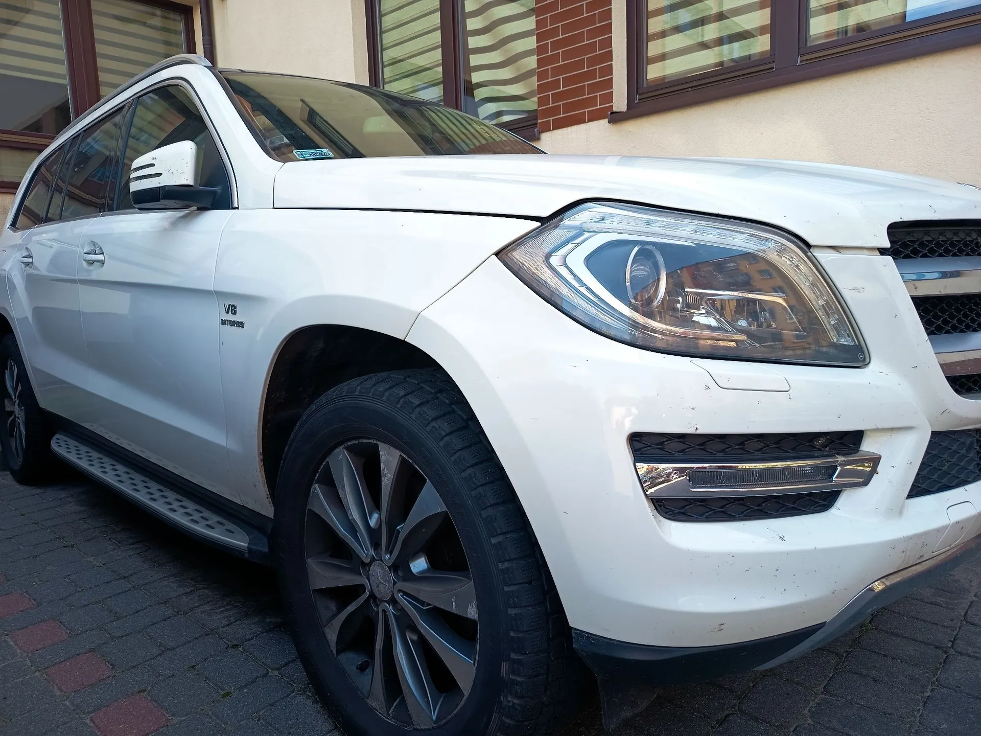 Biały samochód typu SUV zaparkowany na kostce brukowej przed budynkiem. Zdjęcie przedstawia przód i prawy bok pojazdu pod kątem. Widoczny jest reflektor, grill, przednie koło z ciemną felgą oraz nadpis 'V8 BITURBO' na przednim błotniku.