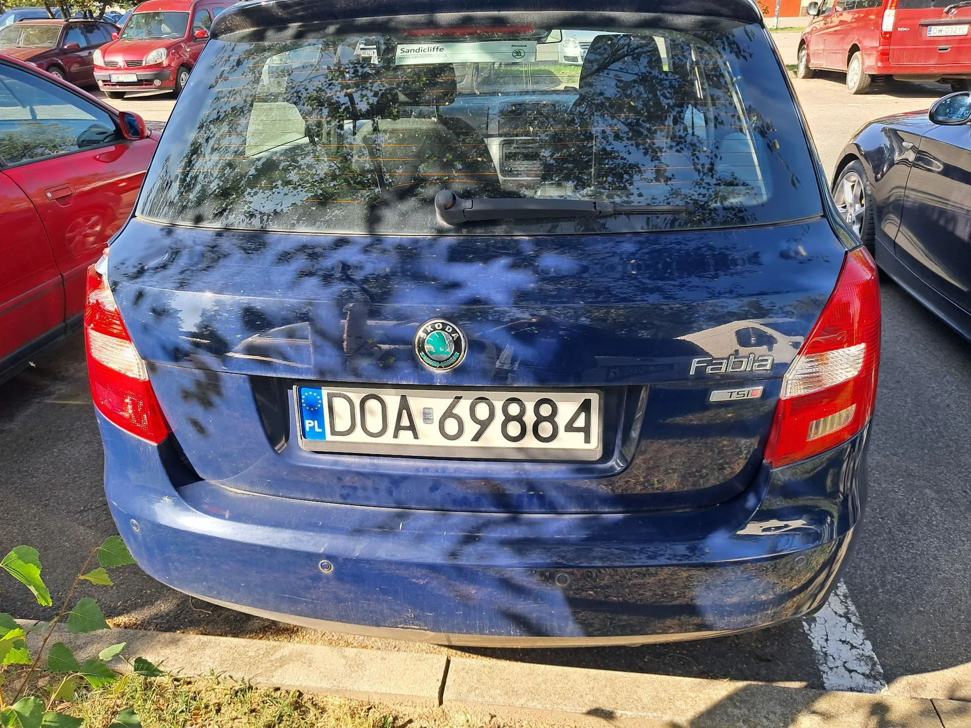 Tylny widok ciemnoniebieskiego samochodu marki Skoda Fablia. Na klapie bagażnika znajduje się logo Skody oraz napis 'Fabia TSI'. Samochód ma polską tablicę rejestracyjną o numerze DOA 69884. Po lewej stronie widoczny jest fragment czerwonego samochodu, a po prawej fragment ciemnego auta. W tle widać inne zaparkowane pojazdy i odbicia drzew w tylnej szybie.