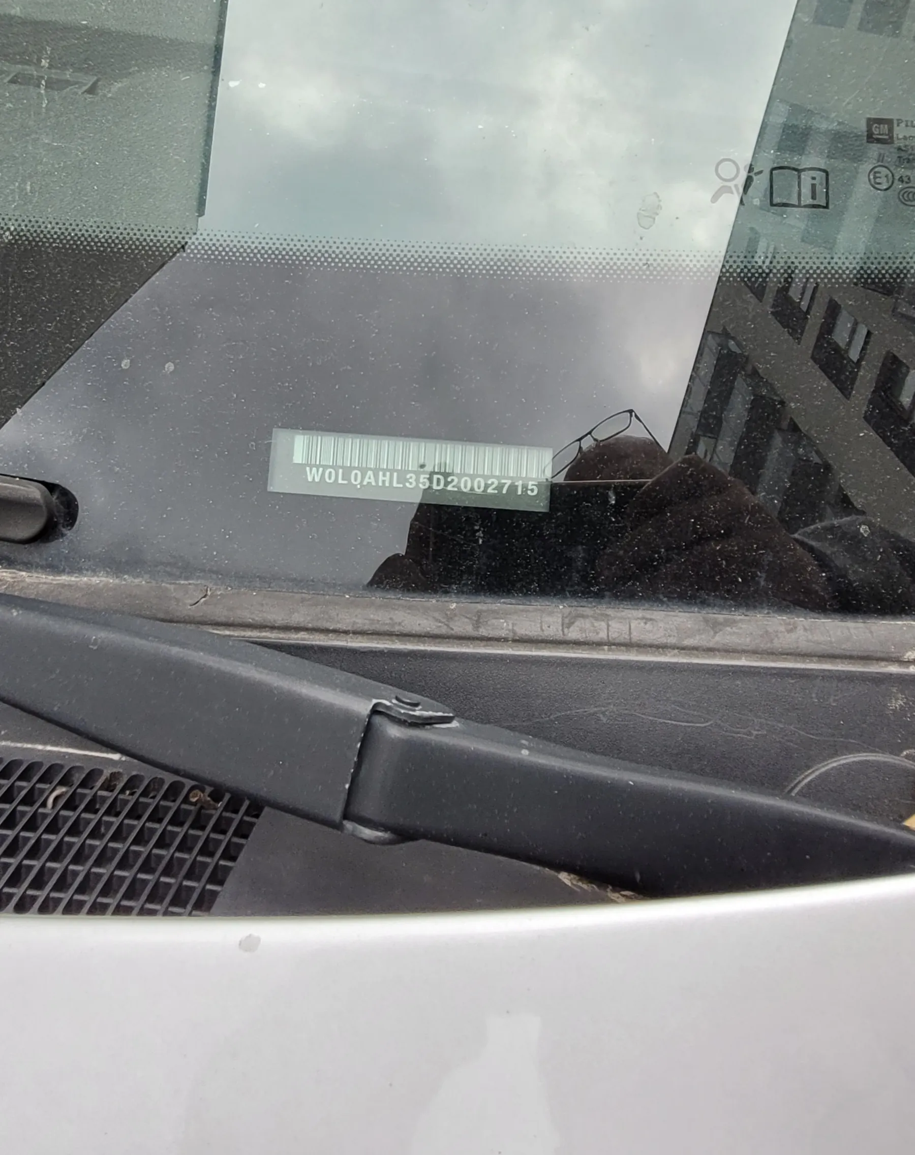 Zbliżenie na przednią szybę samochodu z naklejką zawierającą kod kreskowy i tekst WOLQAH L35D2002715. Widoczny jest odbicie osoby w okularach i ciemnej kurtce, czarny wycieraczka oraz fragment budynku i nieba w tle. Na szybie znajdują się również symbole informacyjne w prawym górnym rogu.