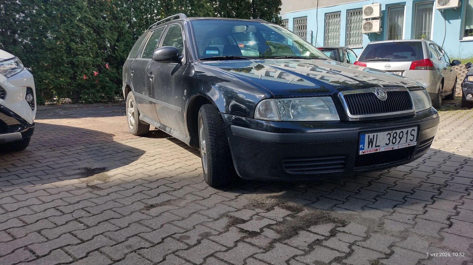 Czarny samochód marki Skoda w nadwoziu kombi zaparkowany na kostce brukowej. Pojazd ma polską tablicę rejestracyjną o numerze WL 3891S. W tle widoczny jest fragment białego samochodu po lewej stronie, srebrny samochód po prawej oraz jasnoniebieski budynek z oknami i jednostkami klimatyzacji na ścianie.
