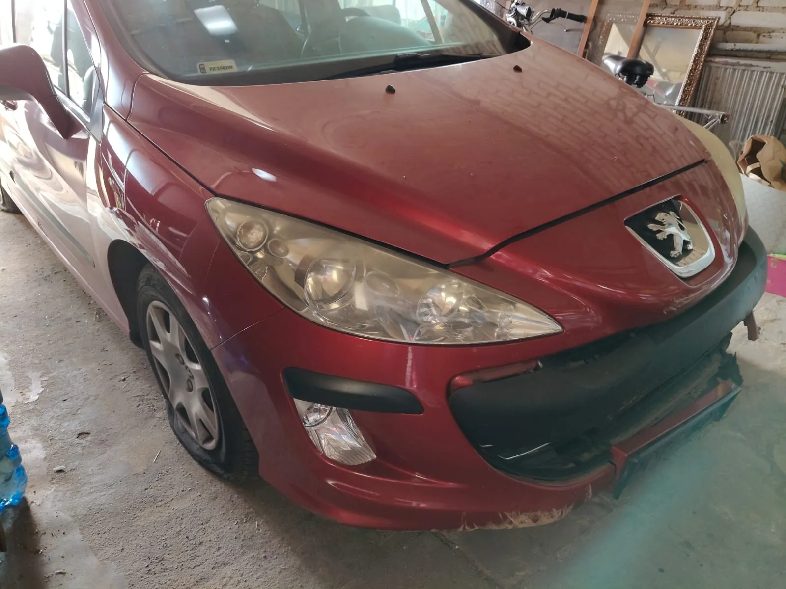 Przednia część czerwonego samochodu marki Peugeot, zaparkowanego w garażu lub warsztacie. Widoczny jest przód pojazdu z logo lwa na masce, reflektory oraz przedni zderzak, który jest częściowo poluzowany i uszkodzony po prawej stronie. Samochód stoi na betonowej posadzce.
