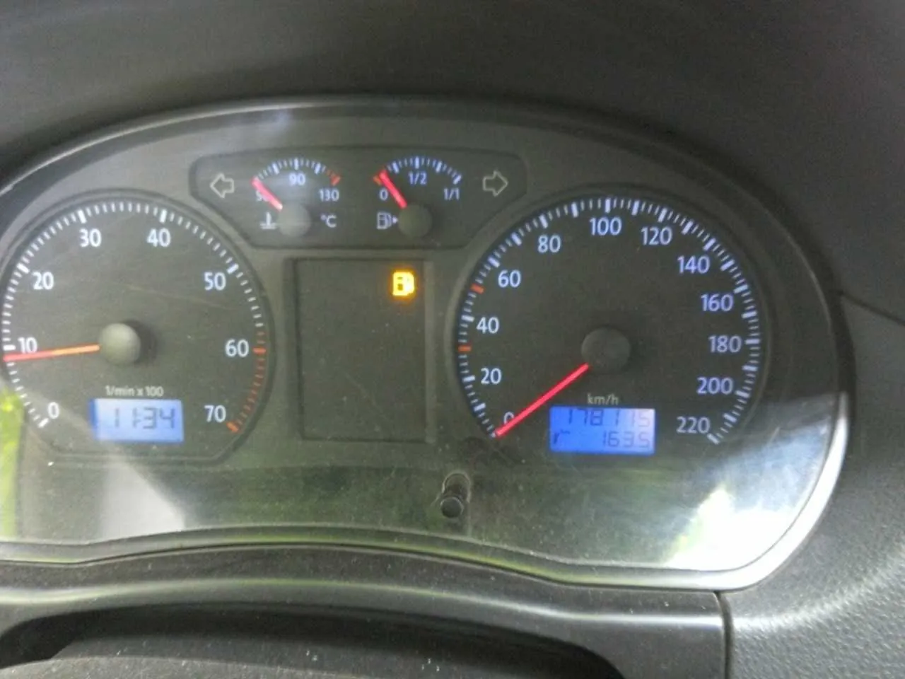 Tablica rozdzielcza samochodu z trzema głównymi wskaźnikami. Po lewej stronie wskaźnik obrotomieru z zakresem 0-70 (1/min x100), na wyświetlaczu cyfrowym wartość 1134. W środku dwa małe wskaźniki: termometr z zakresem 0-130 °C i wskaźnik paliwa z podziałką 0-1/2. Poniżej żarówka ostrzegawcza z literą B. Po prawej stronie wskaźnik prędkości z zakresem 0-220 km/h, na wyświetlaczu cyfrowym wartości 118 (prędkość) i 163,5. Wskaźniki mają niebieskie i czerwone oznaczenia, tło tablicy jest ciemne. Widoczne strzałki kierunkowskazów po lewej i prawej stronie centralnej części.