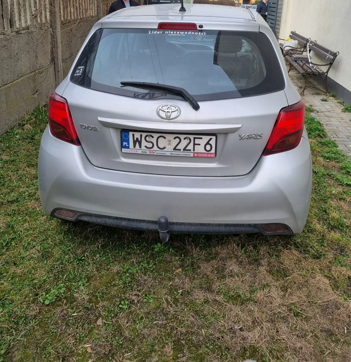 Srebrny samochód Toyota Yaris z tyłu, zaparkowany na trawie. Widoczna tablica rejestracyjna WSC 22F6 z oznaczeniem PL oraz napisem "Lider niezawodności" na tylnej szybie. Po lewej stronie betonowa ściana, po prawej ławka i budynek. Na tablicy rejestracyjnej widoczne dodatkowe napisy: "TOYOTA", "W&J", "Warszawa-Łobelin" oraz numer telefonu. Trawa wokół samochodu jest częściowo sucha.