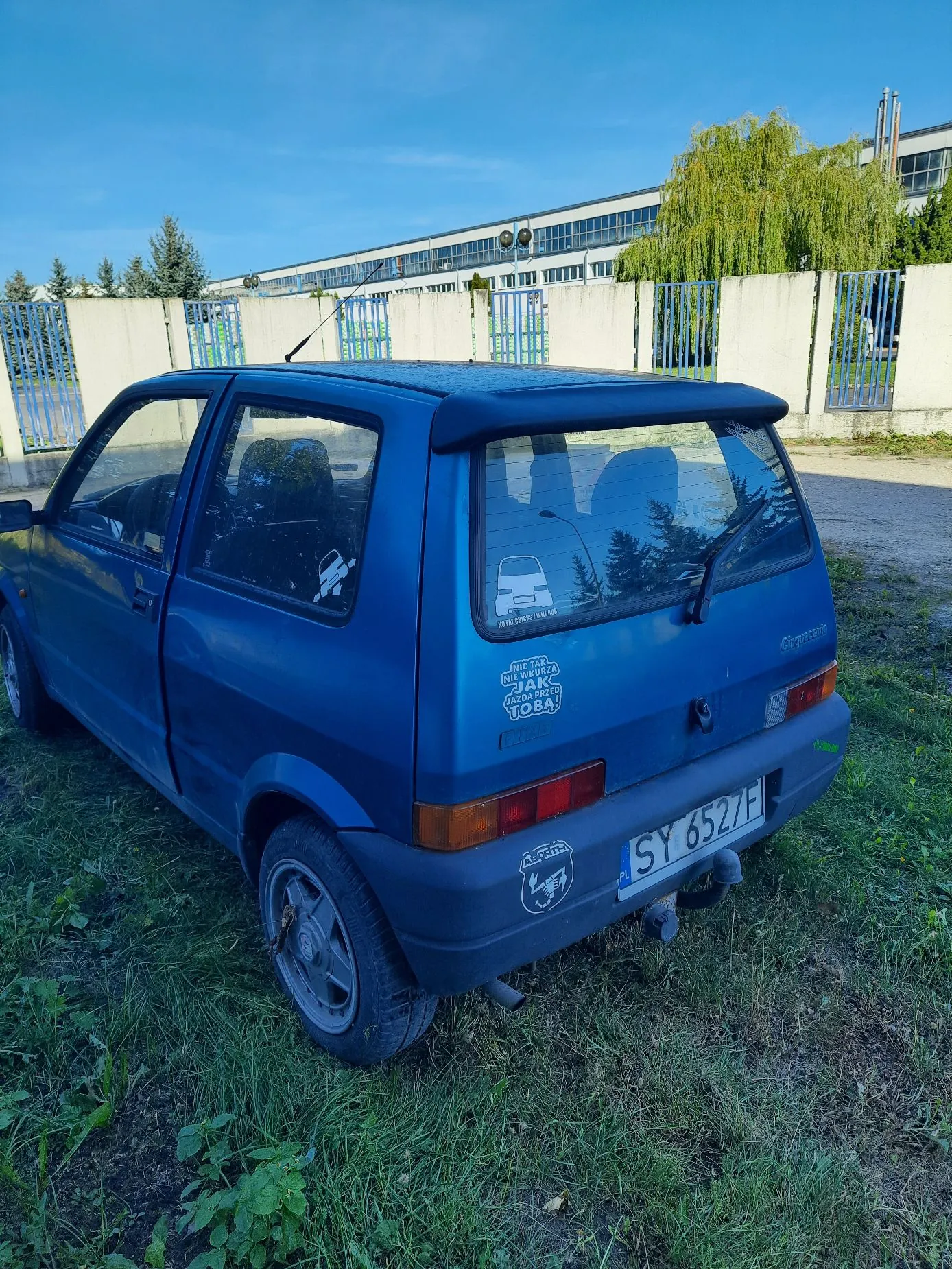 Niebieski samochód osobowy marki Fiat Cinquecento zaparkowany na trawiastym poboczu. Na tylnej szybie znajduje się naklejka z białym samochodem i napisem „NIE ME DREWCY - NIE TEGO”, a poniżej na klapie bagażnika biała naklejka z tekstem „NIC TAK NIE SZKODZI ZIARKU JAKU ZIARNO TOBA!”. Samochód ma polską tablicę rejestracyjną o numerze SY 6527F. W tle widoczny jest biały płot z niebieskimi bramami oraz długi, jasny budynek przemysłowy pod niebieskim niebem.