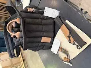 Czarna pikowana kurtka leżąca na ciemnej powierzchni. Na kurtce widoczne są białe etykiety z nieczytelnym tekstem oraz brązowa etykieta. Obok kurtki znajduje się beżowa torba z brązowym paskiem i metalowym zamkiem oraz brązowy pasek z metalową klamrą. W tle widoczne są pudełka i drewniana konstrukcja.