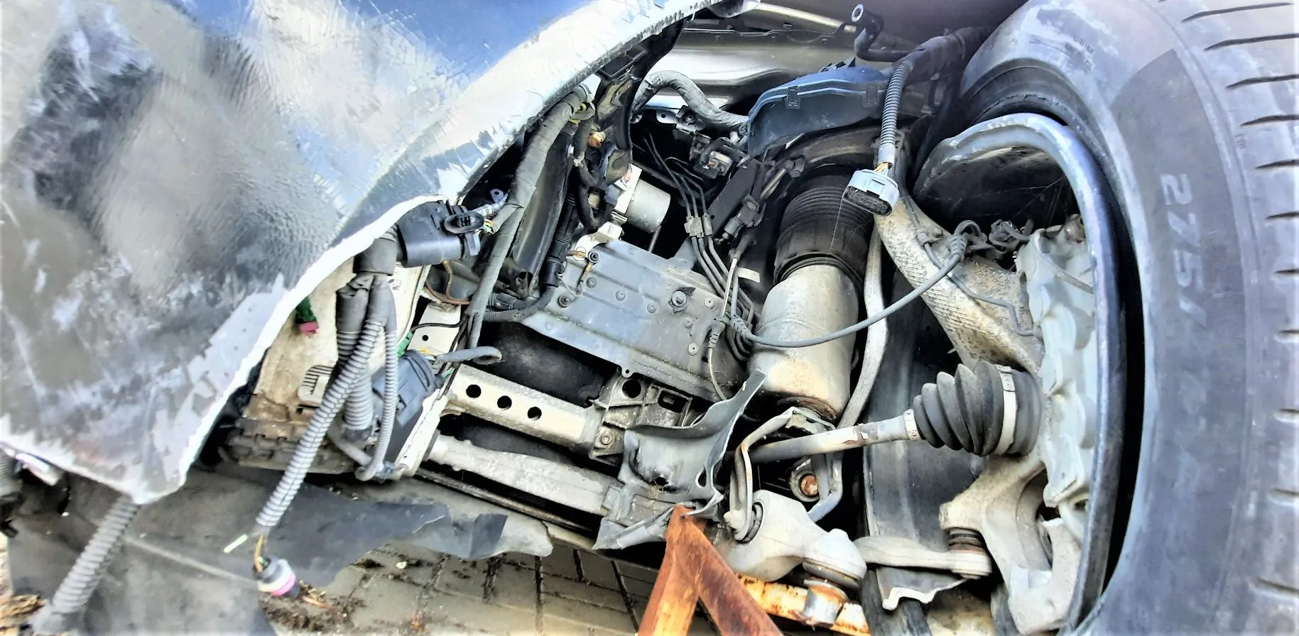 Zbliżenie na uszkodzony przód samochodu z widocznymi elementami zawieszenia, silnika i nadwozia. Widoczna jest opona z napisem '275/40', amortyzator, przewody elektryczne w peszlach oraz zgnieciona blacha błotnika. Pojazd spoczywa na podłożu, pod spodem widoczny jest metalowy element wspierający.
