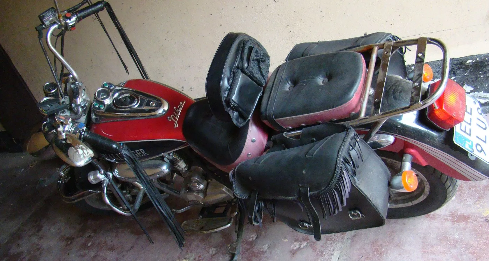 Czerwony motocykl typu cruiser zaparkowany w pomieszczeniu. Pojazd posiada czarne skórzane siedzenia, dwa duże czarne kufry boczne z frędzlami oraz metalowy bagażnik z tyłu. Widoczny jest chromowany kierownica, reflektor oraz fragment tablicy rejestracyjnej z tyłu.