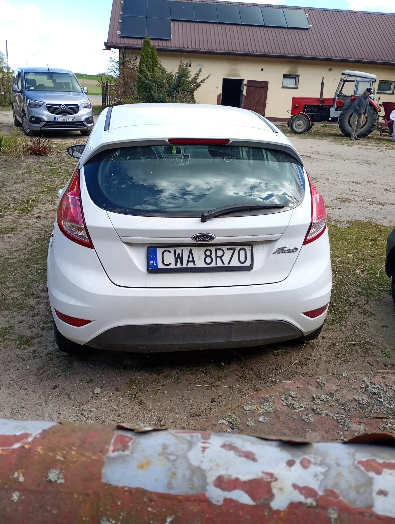 Biały samochód Ford Fiesta z tablicą rejestracyjną CWA 8R70 (PL) widziany od tyłu. W tle srebrny samochód Opel z tablicą CB 617LW, czerwony traktor, żółty budynek z brązowym dachem i panelami słonecznymi oraz osoba stojąca obok traktora. Na pierwszym planie rdzewiejąca metalowa bariera. Tło stanowi podwórze z ziemią i trawą.