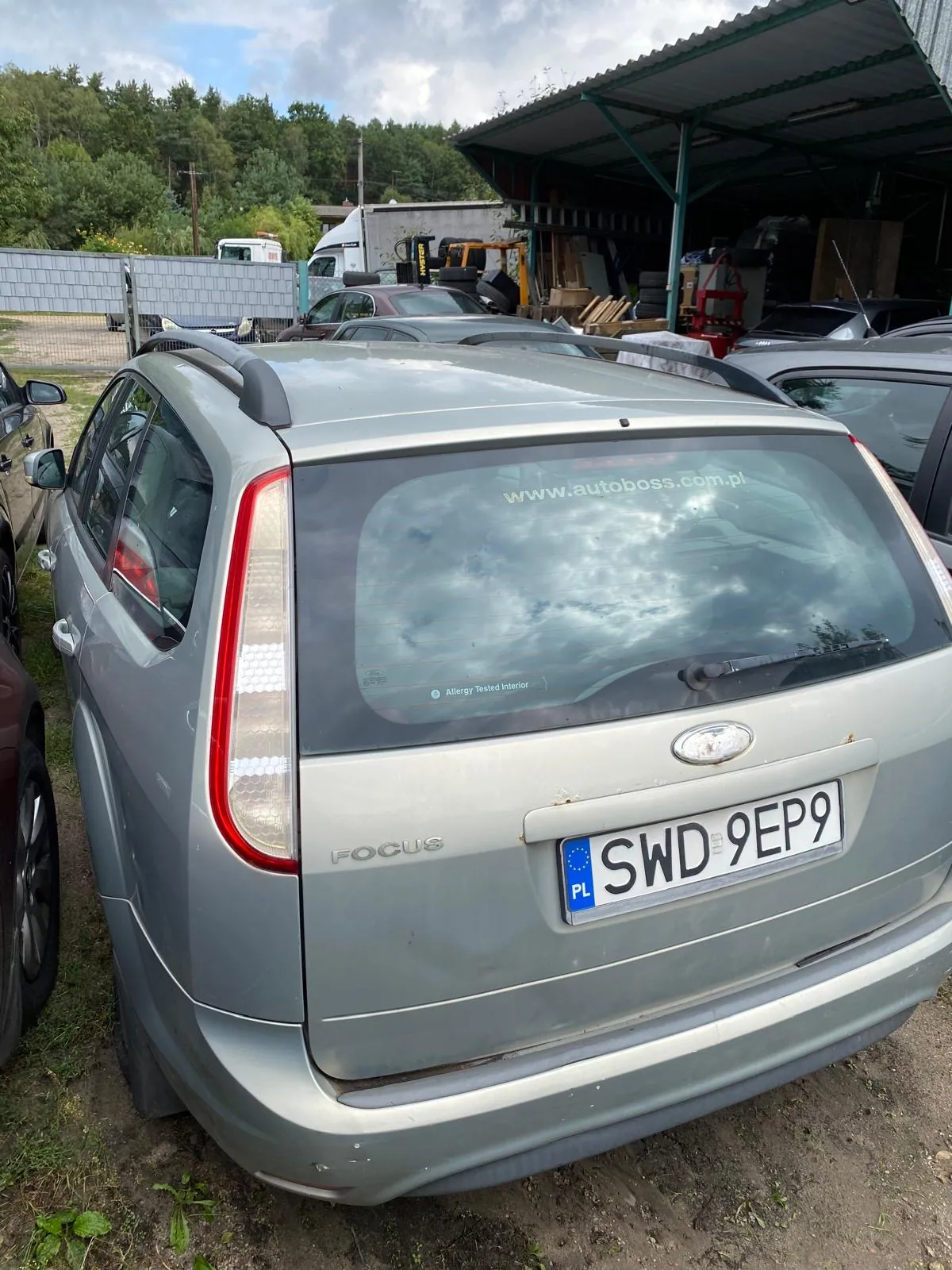 Tylny widok srebrnego samochodu marki Ford Focus w wersji kombi. Na tylnej szybie znajduje się napis 'www.autoboss.com.pl'. Samochód posiada polską tablicę rejestracyjną o numerze SWD 9EP9. Pojazd stoi na piaszczystym podłożu na placu z innymi samochodami i zadaszoną wiatą w tle.