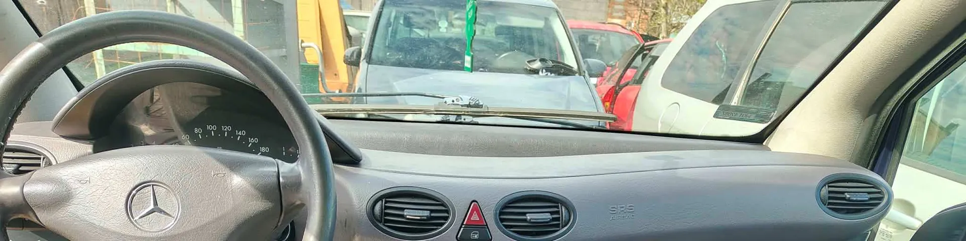 Wnętrze samochodu z widoczną kierownicą z logo Mercedes-Benz, tablicą przyrządów z prędkościomierzem do 180 km/h, wentylacjami i napisem SRS AIRBAG. Przez przednią szybę widać inne pojazdy: biały van z zielonym znaczkiem na szybie, czerwony samochód oraz żółte maszyny budowlane na tle. Wnętrze pojazdu ma szary wykończenie, a na desce rozdzielczej znajduje się czerwony trójkątny symbol ostrzegawczy.