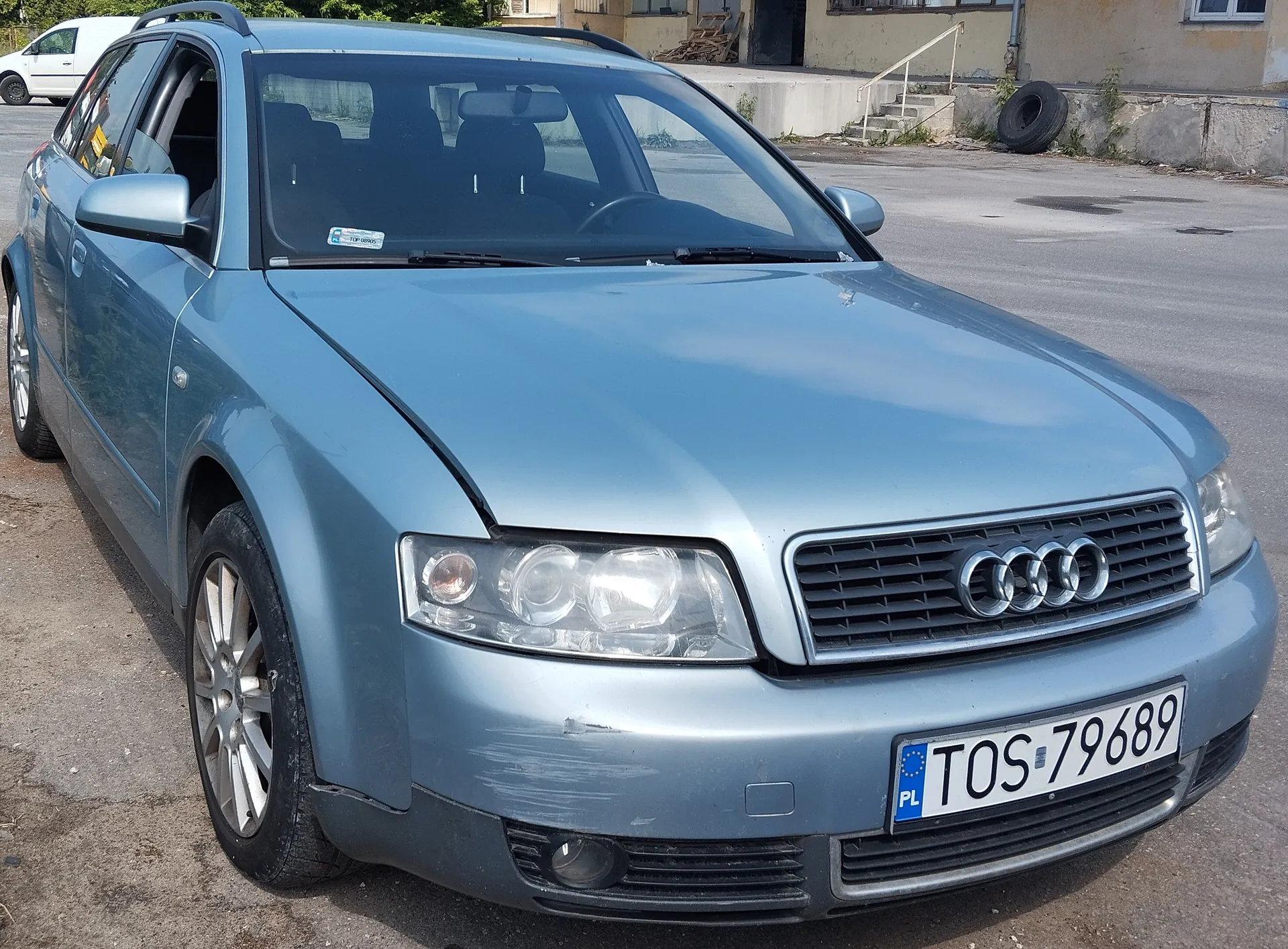 Srebrny samochód Audi typu kombi zaparkowany na asfaltowym placu. Widoczna tablica rejestracyjna z polskim kodem 'PL' i numerem 'TOS-79689'. W tle budynek z betonowymi schodami, oponą opartą o mur oraz biały van. Na przedniej szybie naklejka.