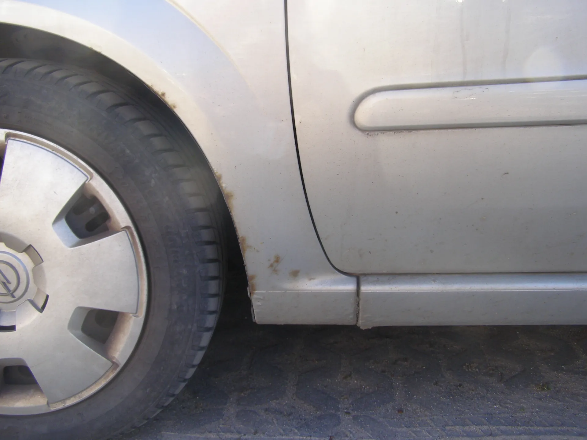 Zbliżenie na przednie koło i fragment boku srebrnego samochodu. Widoczna opona z metalowym felgiem z logo, brud na karoserii oraz rdza w okolicy otworu kołowego. Samochód stoi na asfaltowej powierzchni z widocznymi śladami opon.