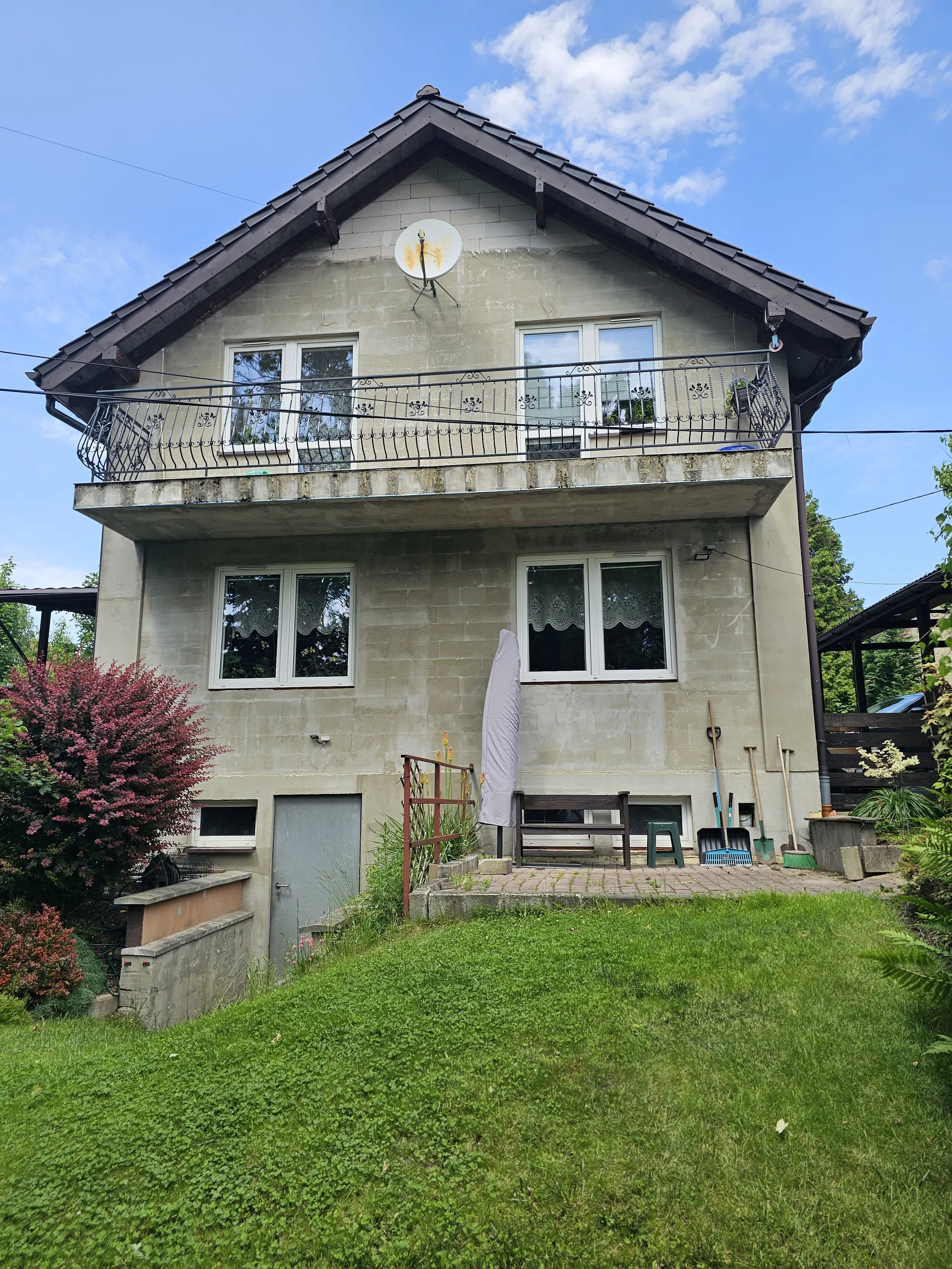 Dwupiętrowy dom o beżowej elewacji z ciemnym dachem. Na ścianie satelita, balkony z czarnymi metalowymi balustradami. Okna z białymi ramami i koronkowymi zasłonami. Przed domem zielona trawa, czerwony krzew, składany parasol, narzędzia ogrodnicze (łopaty, grabie), kamienna ścieżka i schody. Niebo z białymi chmurami.