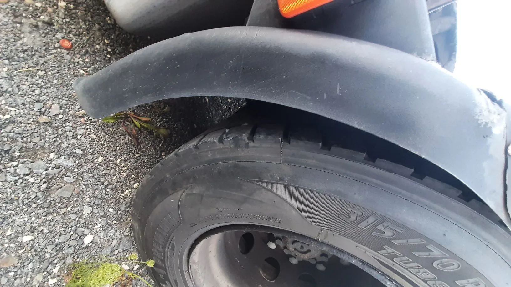 Zbliżenie na tylne koło pojazdu z czarnym, plastikowym błotnikiem nad oponą. Opona ma widoczny napis '315/70 R'. Pojazd stoi na szutrowym podłożu.