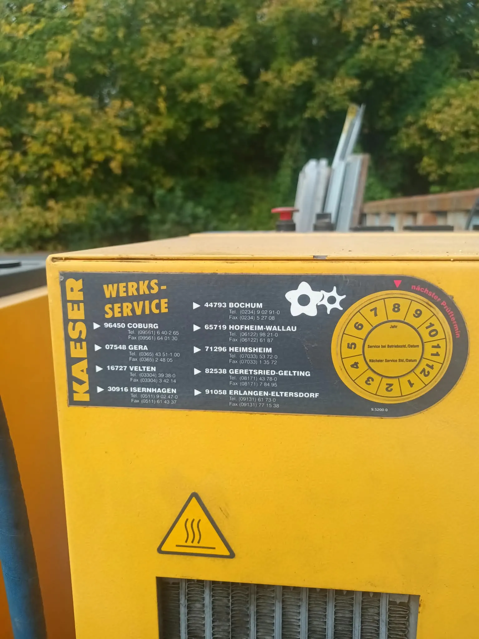 Zbliżenie na żółtą obudowę urządzenia marki KAESER z naklejką 'WERKS-SERVICE'. Na naklejce znajduje się lista lokalizacji serwisowych w Niemczech wraz z numerami telefonów i faksów, m.in.: Coburg, Bochum, Hofheim-Wallau, Gera, Heimsheim, Velten, Geretsried-Gelting, Isernhagen oraz Eltersdorf. Po prawej stronie znajduje się okrągły wykres serwisowy z numerami od 1 do 12 i polami do wpisania daty. Poniżej naklejki widoczny jest trójkątny znak ostrzegawczy przed gorącą powierzchnią.