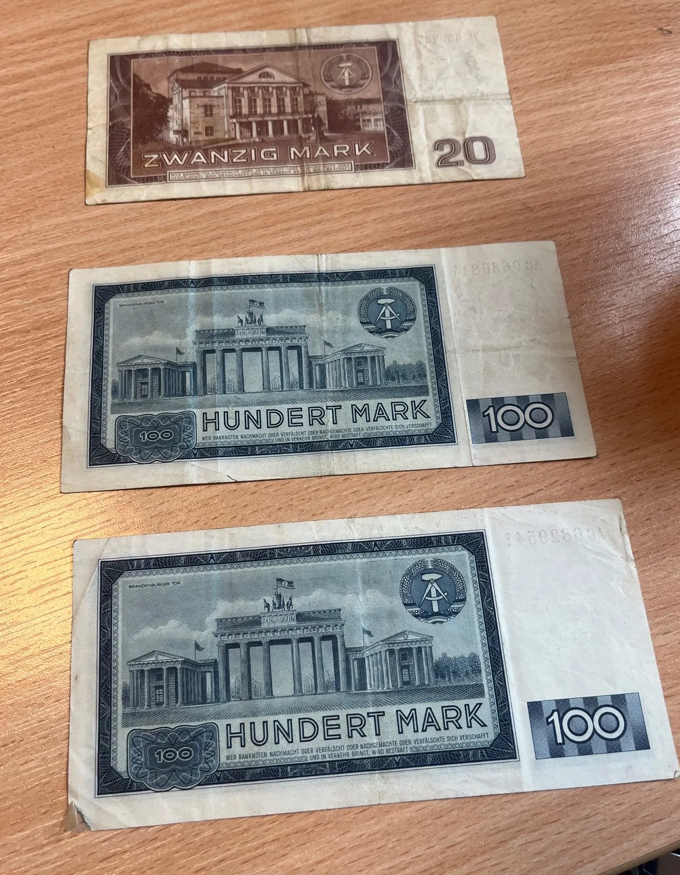 Trzy stare niemieckie banknoty leżące na drewnianym blacie. Górny banknot jest brązowy z nominałem 20 i napisem 'ZWANZIG MARK'. Dwa dolne banknoty są niebieskie z nominałem 100 i napisem 'HUNDERT MARK'. Na niebieskich banknotach widnieje ilustracja Bramy Brandenburskiej w Berlinie oraz tekst: 'WER BANKNOTEN NACHMACHT ODER VERFÄLSCHT ODER NACHGEMACHTE ODER VERFÄLSCHTE SICH VERSCHAFFT, VERWENDET UND IN BESITZ NAHÄBT BRINGT, W. RD BESTRAFT'.