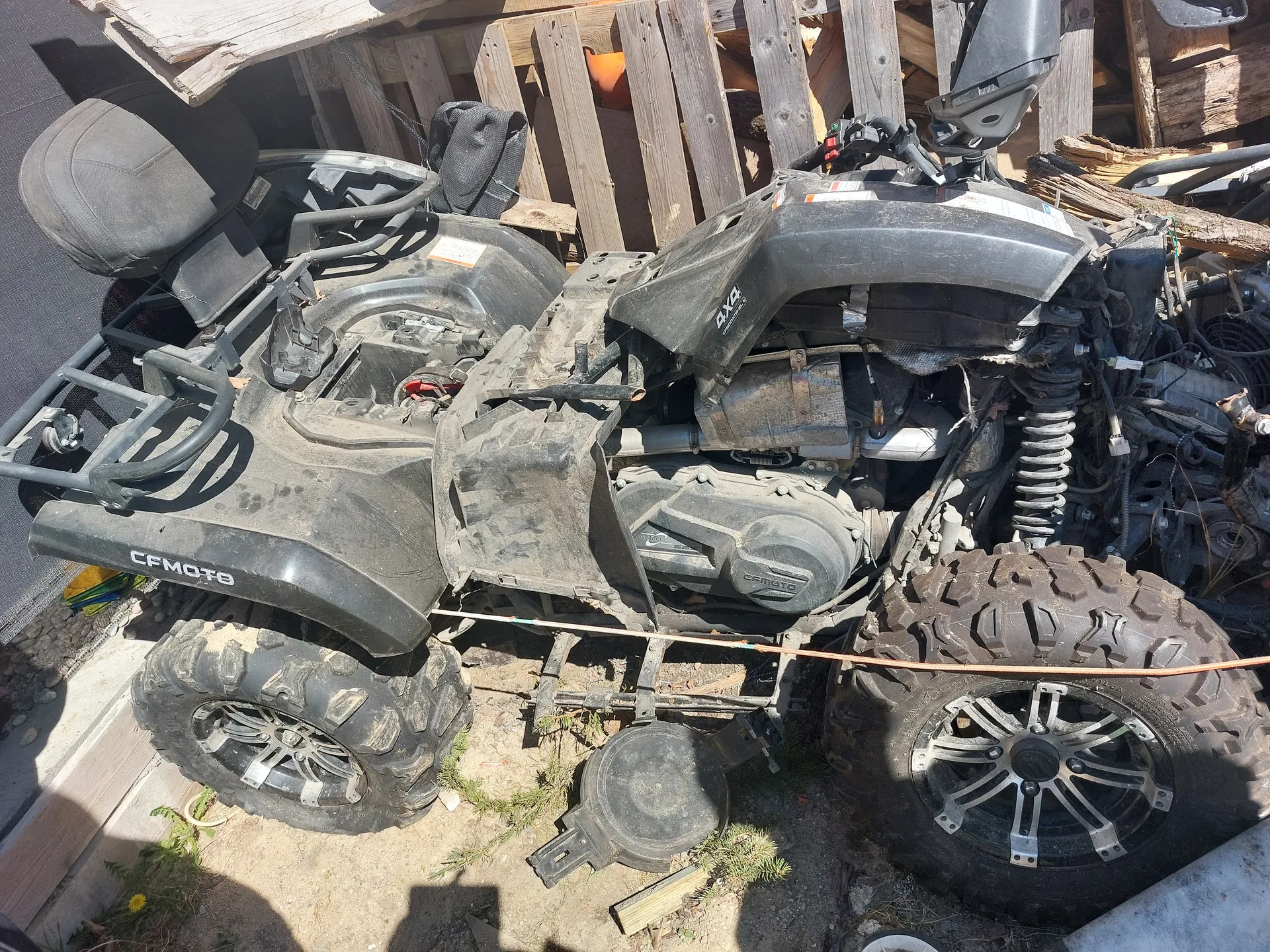 Czarny quad marki CFMOTO w stanie częściowej rozbiórki, z widocznymi zanieczyszczonymi oponami, silnikiem i elementami mechanizmu. Pojazd znajduje się na brudnej powierzchni ziemnej z fragmentami trawy, w tle drewniane deski i różne części mechaniczne. Na boku kadłuba widoczny napis CFMOTO.