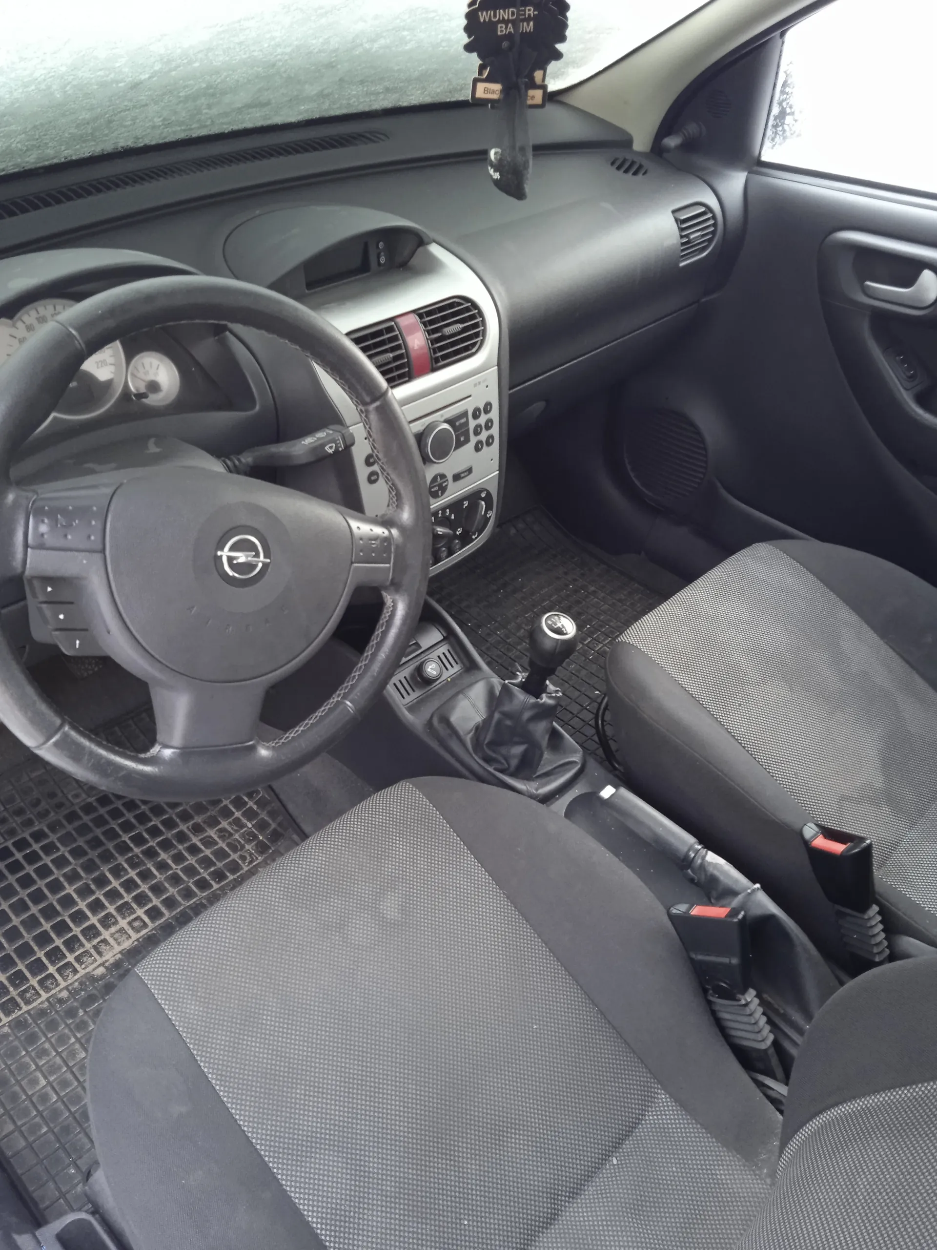 Wnętrze samochodu z widokiem na kierownicę z logo Opel, deska rozdzielcza z przyciskami i wentylacją, dźwignia zmiany biegów, czarne siedzenia z szarym wzorem oraz dywaniki podłogowe. Na lusterku wstecznym wisząca ozdoba z napisem "WUNDER-BAUM" i małą tabliczką "Black". W tle widoczne liczniki prędkości i obrotomierz.