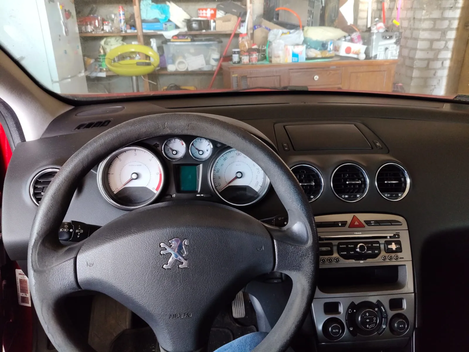 Widok z wnętrza samochodu marki Peugeot, skupiony na kierownicy i desce rozdzielczej. Na kierownicy widoczne jest srebrne logo lwa oraz napis AIRBAG. Przed kierownicą znajduje się zestaw wskaźników z prędkościomierzem, obrotomierzem i mniejszymi zegarami. Po prawej stronie widać trzy okrągłe nawiewy, panel radia oraz sterowanie klimatyzacją. Przez przednią szybę widoczny jest nieuporządkowany garaż lub warsztat z półkami, przedmiotami i meblami.