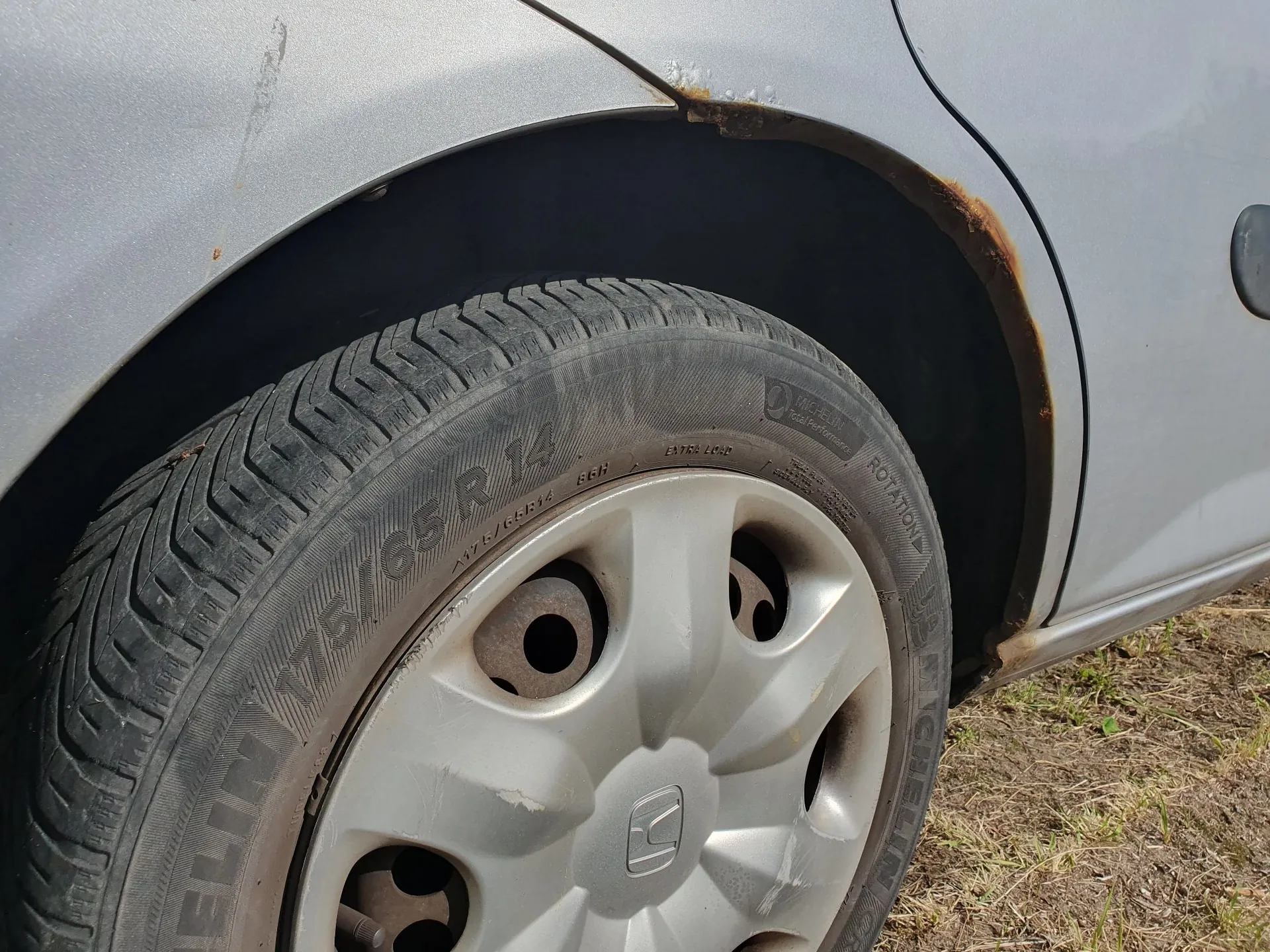 Zbliżenie na przednie koło srebrnego samochodu. Widoczna jest opona z napisem 'MICHELIN 175/65 R 14' oraz srebrna felga z logo marki Hyundai. Nad nadkolem samochodu widoczne są wyraźne ślady korozji i rdzy na lakierze.