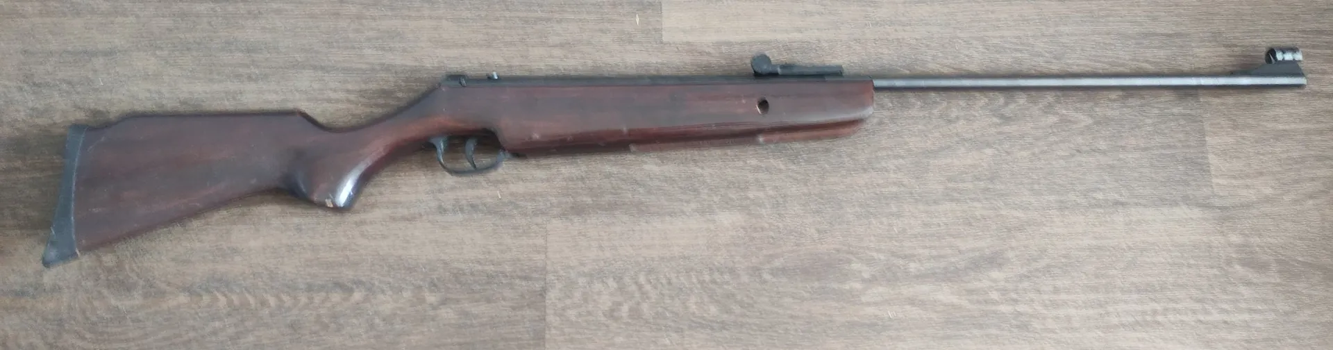 Karabin z drewnianą kolbą i ciemną lufą, leżący poziomo na drewnianej podłodze.