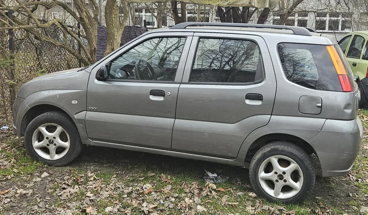 Szary samochód osobowy typu hatchback zaparkowany na trawie z suchymi liśćmi. Pojazd jest pokazany z profilu od strony kierowcy. W tle widoczne są bezlistne drzewa, metalowy płot oraz fragment jasnego budynku z oknami.
