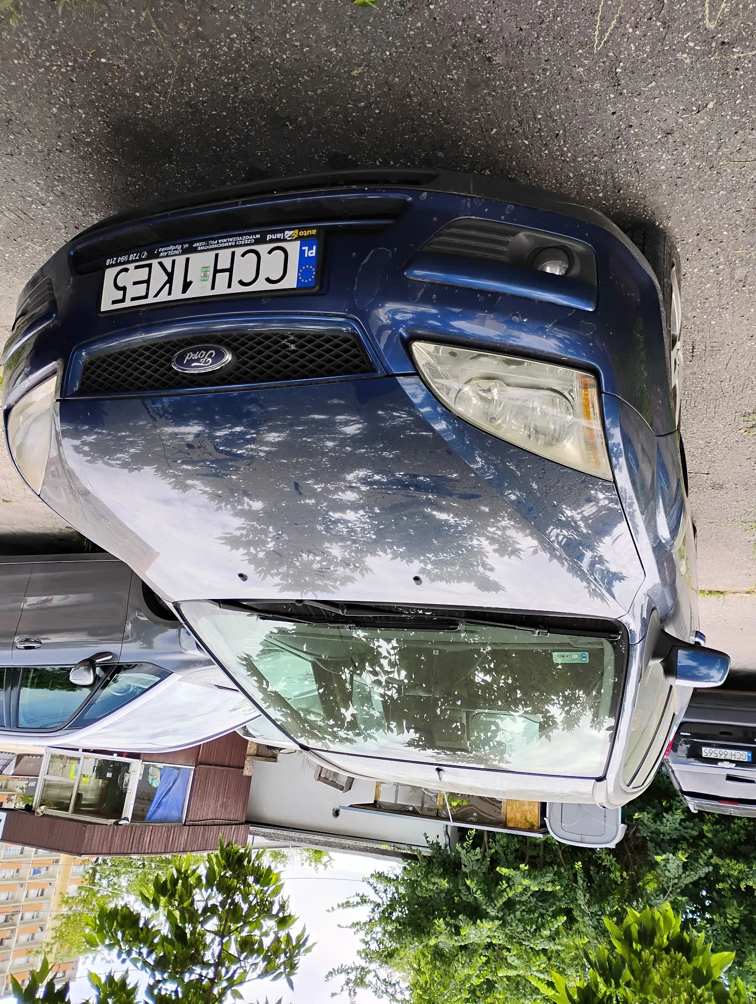 Niebieski samochód marki Ford zaparkowany na asfaltowej nawierzchni. Widoczna jest przednia część pojazdu z tablicą rejestracyjną o numerze CCH 1KE5. W tle widać fragmenty innych samochodów, zielone drzewa oraz fragment budynku mieszkalnego.