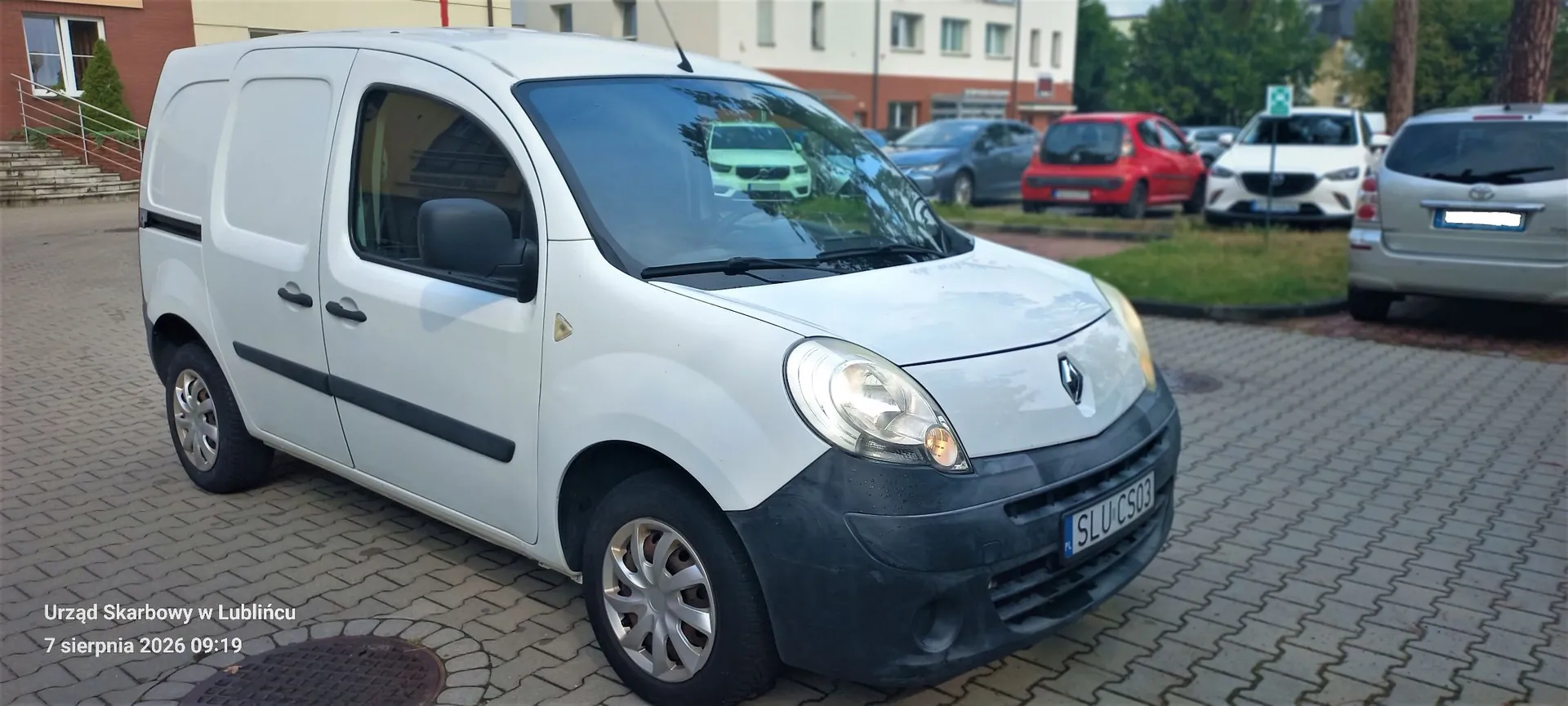 Biały samochód dostawczy marki Renault zaparkowany na kostce brukowej. Pojazd jest ustawiony pod kątem, widoczny z przodu i z lewej strony. Na tablicy rejestracyjnej widnieje napis SLU CS03. W tle znajdują się inne zaparkowane samochody w kolorach niebieskim, czerwonym, białym i srebrnym oraz fragmenty budynków. W lewym dolnym rogu obrazu znajduje się tekst: 'Urząd Skarbowy w Lublińcu 7 sierpnia 2026 09:19'.