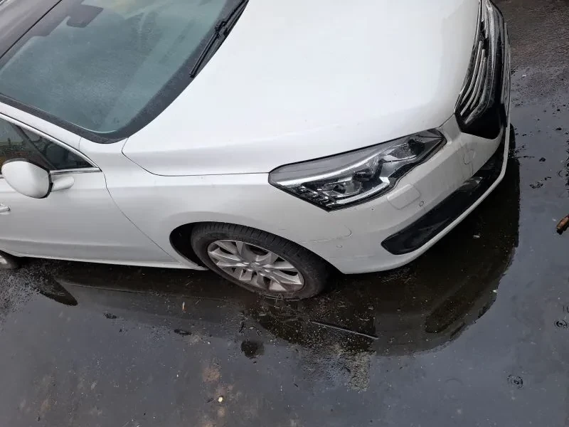 Biały samochód widziany z góry, pokazujący przód pojazdu w tym reflektor, zderzak i koło przednie. Samochód stoi na mokrym asfalcie z kałużami i drobnymi śmieci. Tło stanowi wilgotna powierzchnia drogi bez widocznego tekstu.