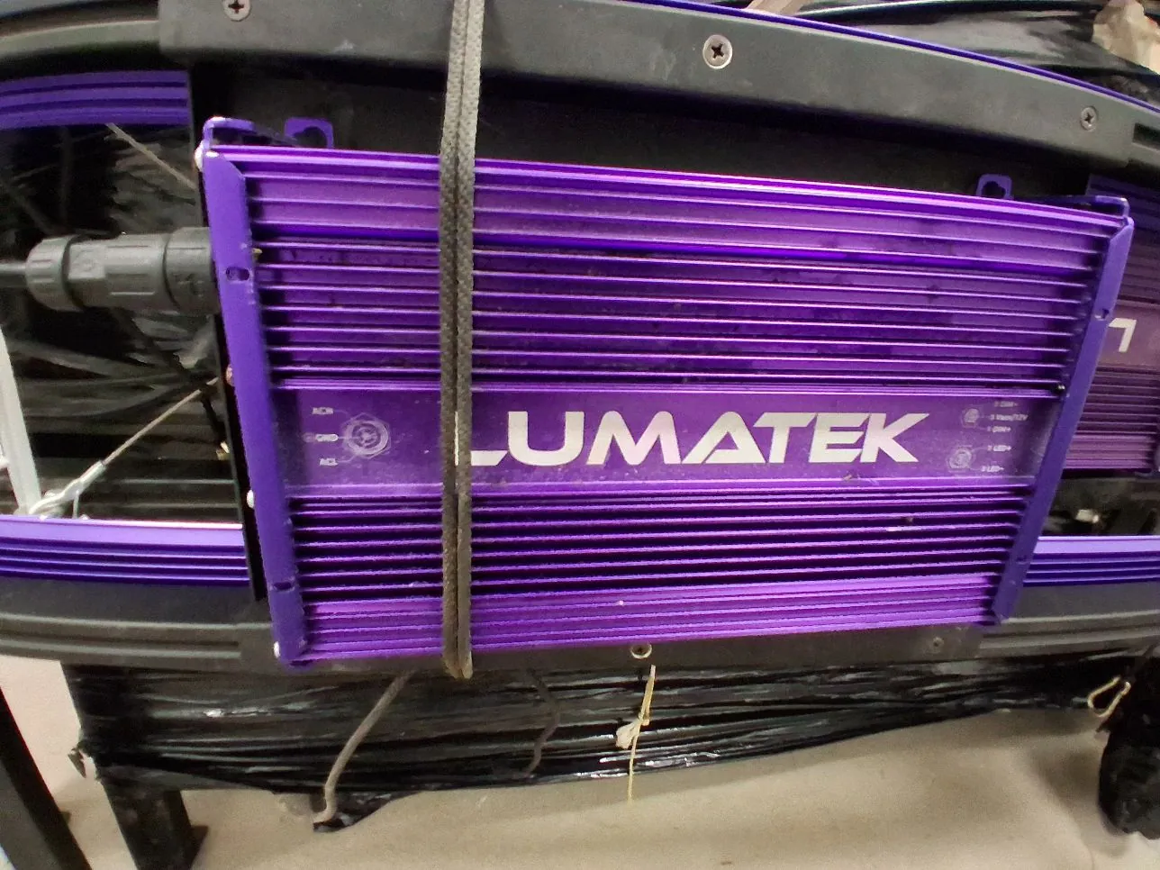 Zbliżenie na fioletowy, prążkowany radiator z białym napisem LUMATEK na środku. Urządzenie jest przymocowane do czarnej konstrukcji i częściowo przebiega przez nie szary pasek. Po lewej stronie widoczne jest czarne złącze kablowe.