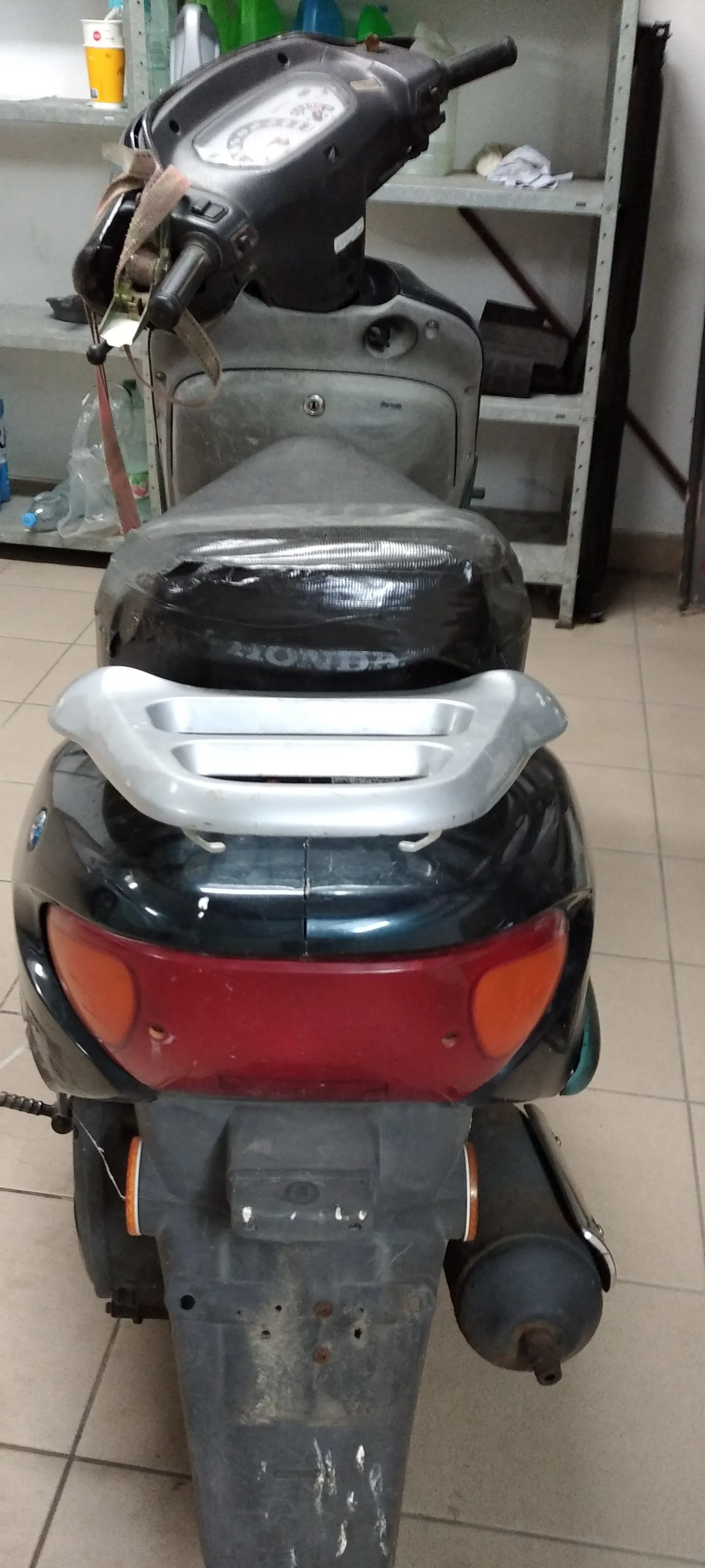 Widok od tyłu na czarny skuter marki Honda. Pojazd posiada srebrny bagażnik nad tylnym światłem, które jest czerwone z pomarańczowymi kierunkowskami po bokach. Skuter stoi w pomieszczeniu na kafelkach, w tle widoczny jest metalowy regał z różnymi przedmiotami.