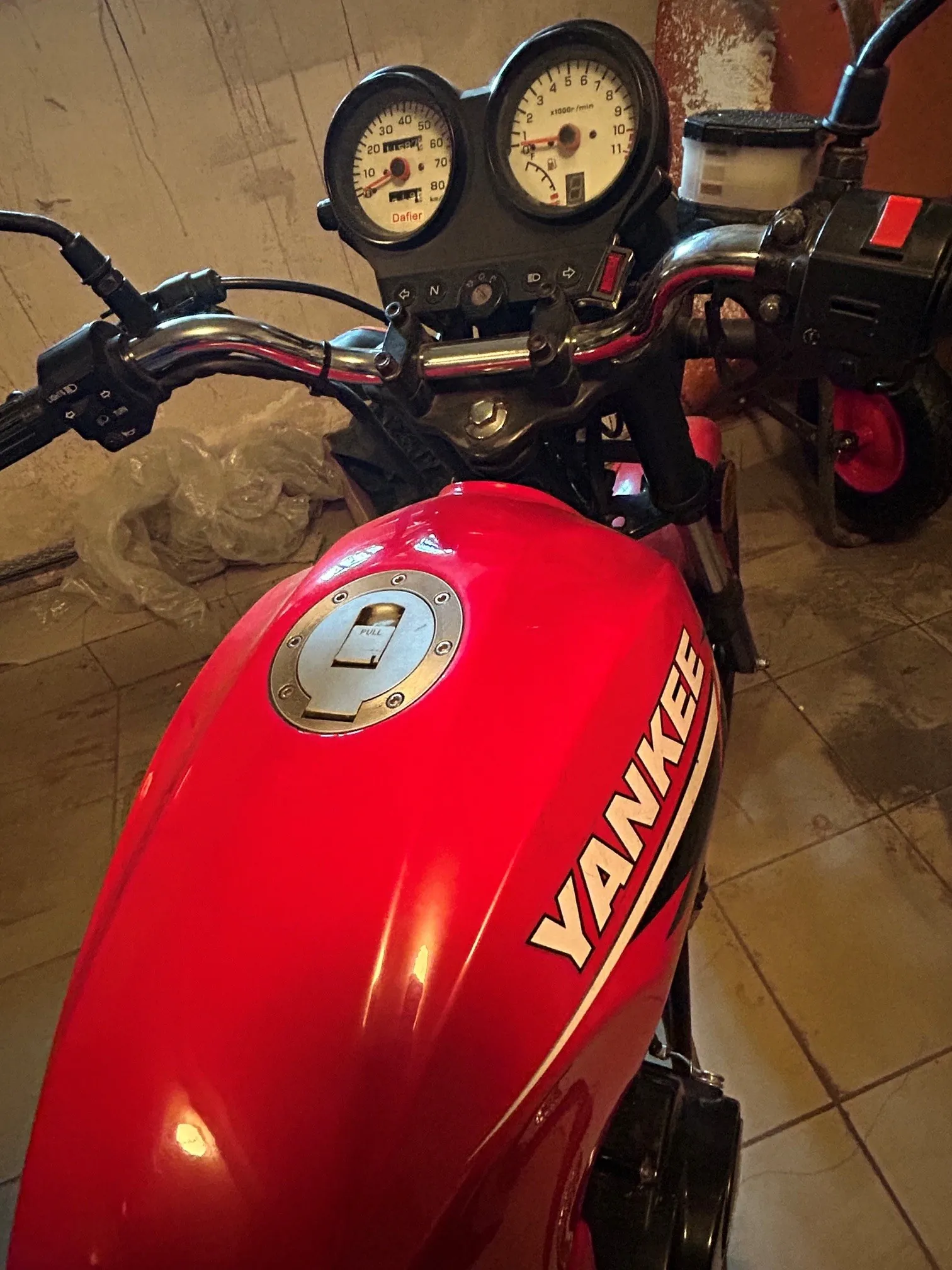 Zbliżenie na przednią część czerwonego motocykla. Widoczny jest czerwony bak z białym napisem 'YANKEE' oraz srebrną zakrętką wlewu paliwa. Powyżej znajduje się kierownica z manetkami oraz dwa okrągłe wskaźniki: prędkościomierz z napisem 'Dafier' oraz obrotomierz.
