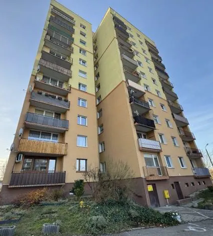 Wysoki blok mieszkalny z dwoma sekcjami: lewa o wysokości około ośmiu pięter w kolorze beżowym z żółtym akcentem, prawa wyższa (około dziesięciu pięter) w kolorze żółtym. Budowla posiada liczne balkony z metalowymi lub szklanymi balustradami. Na parterze widoczne są krzewy, trawnik oraz ścieżka prowadząca do wejścia. W tle jasne niebo bez chmur. Przy wejściu znajdują się żółte tablice informacyjne, ale tekst na nich nie jest czytelny.