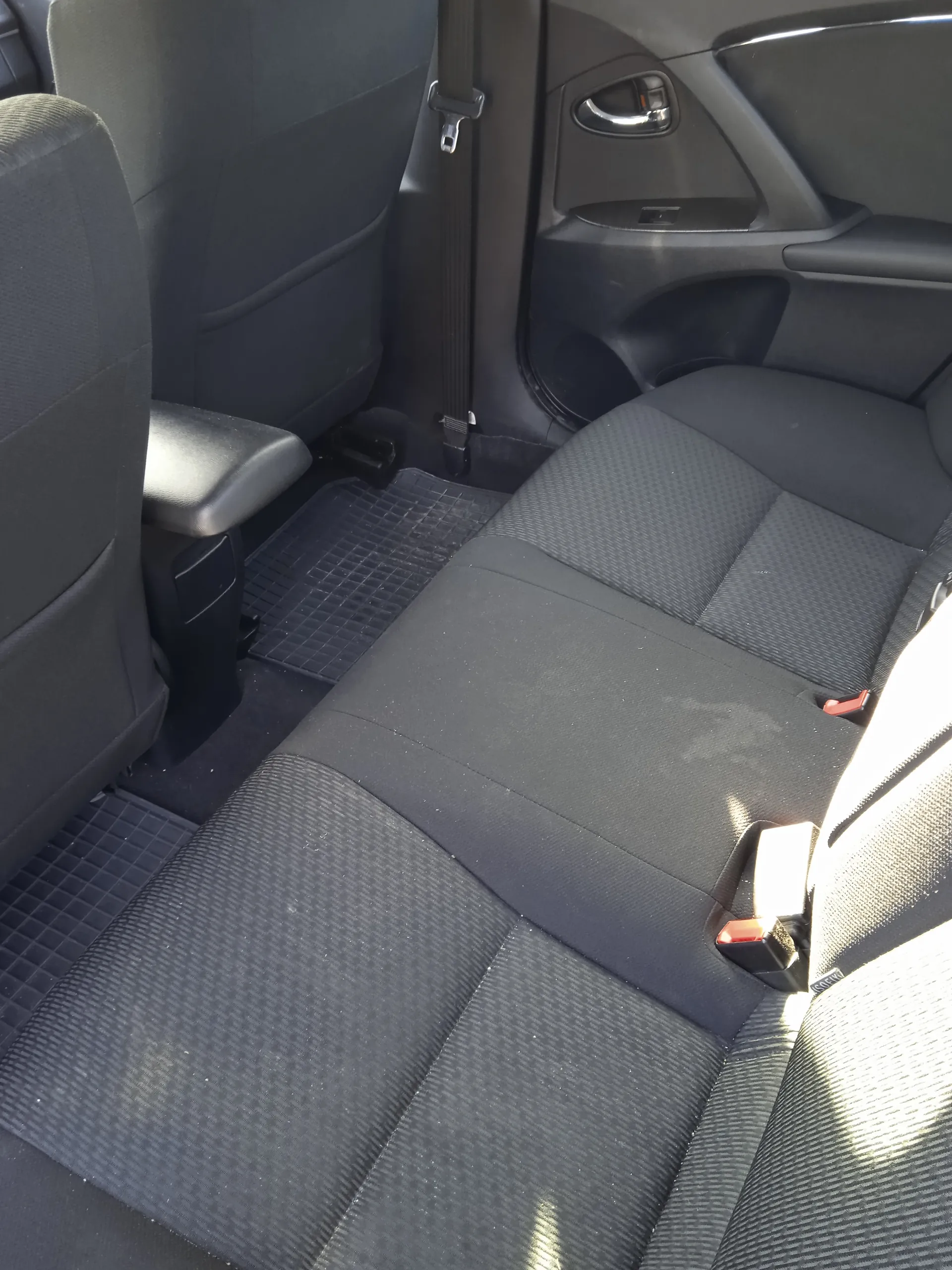 Wnętrze samochodu z widokiem na czarne tylne siedzenia z teksturą, czarne maty podłogowe, pas bezpieczeństwa oraz fragment drzwi z uchwytem i panelami. Siedzenia i elementy są ustawione w standardowej pozycji.